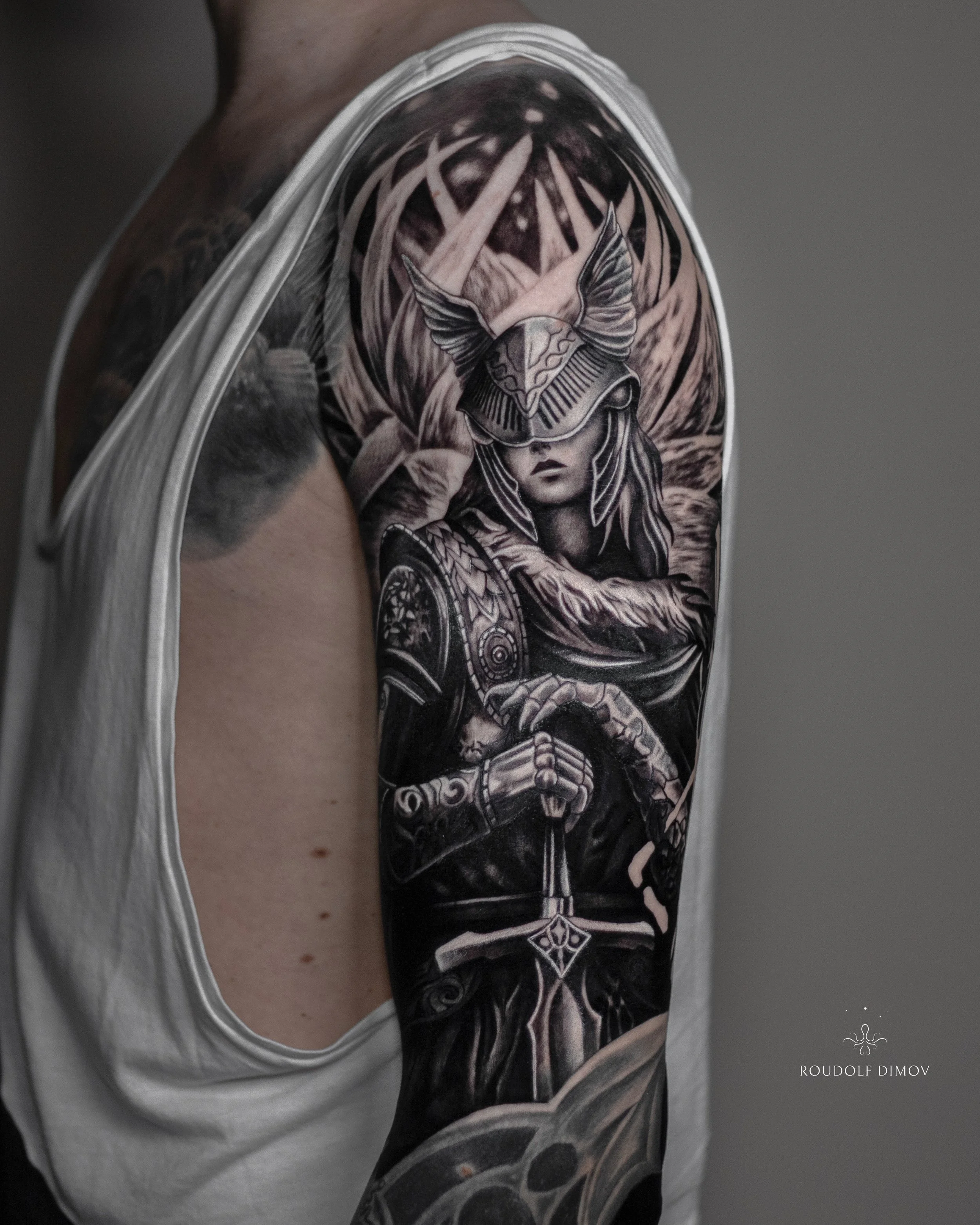 Valkyrie warrior tattoo on the upper arm in black and grey realism, featuring Norse mythology armour, helmet details, and a sword. Tattoo artist Roudolf Dimov, London.