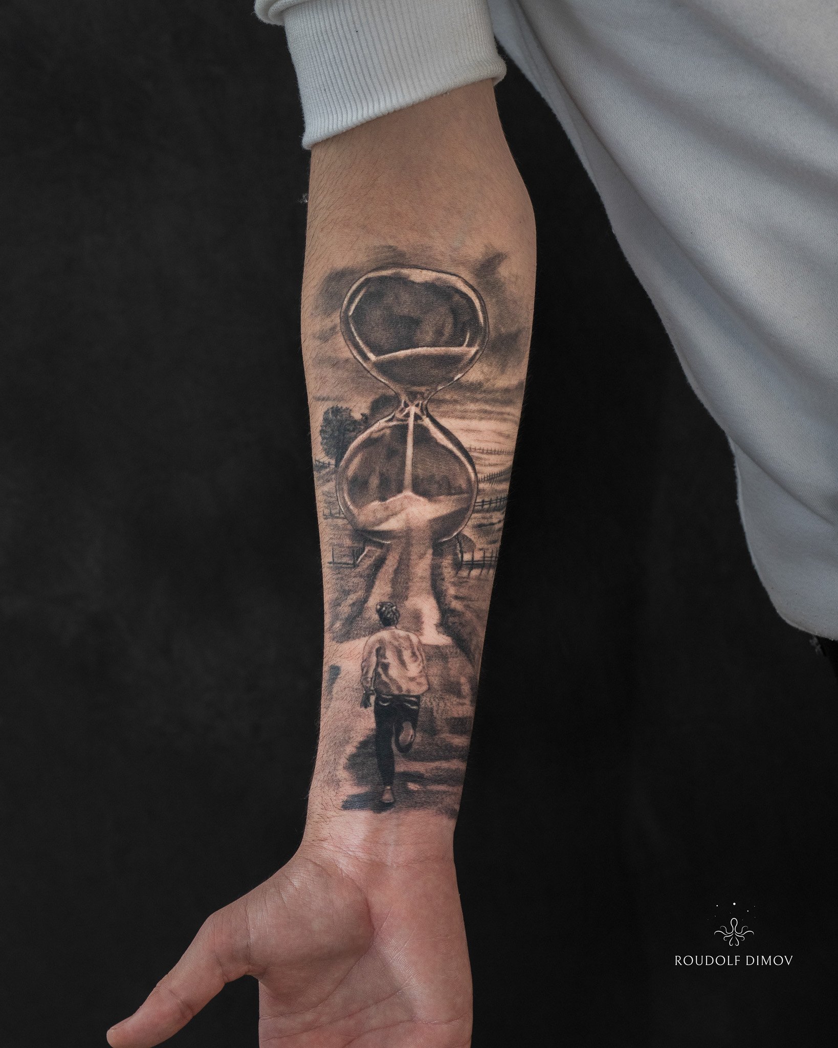 Black & grey realism timepiece tattoo on the outer forearm, featuring an hourglass and running man symbolism. Tattoo by Roudolf Dimov in London.