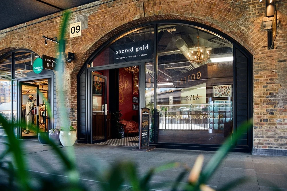 Exterior view of Sacred Gold tattoo, piercing, and jewelry shop with large glass windows and brick archway, overlooking an indoor shopping mall.