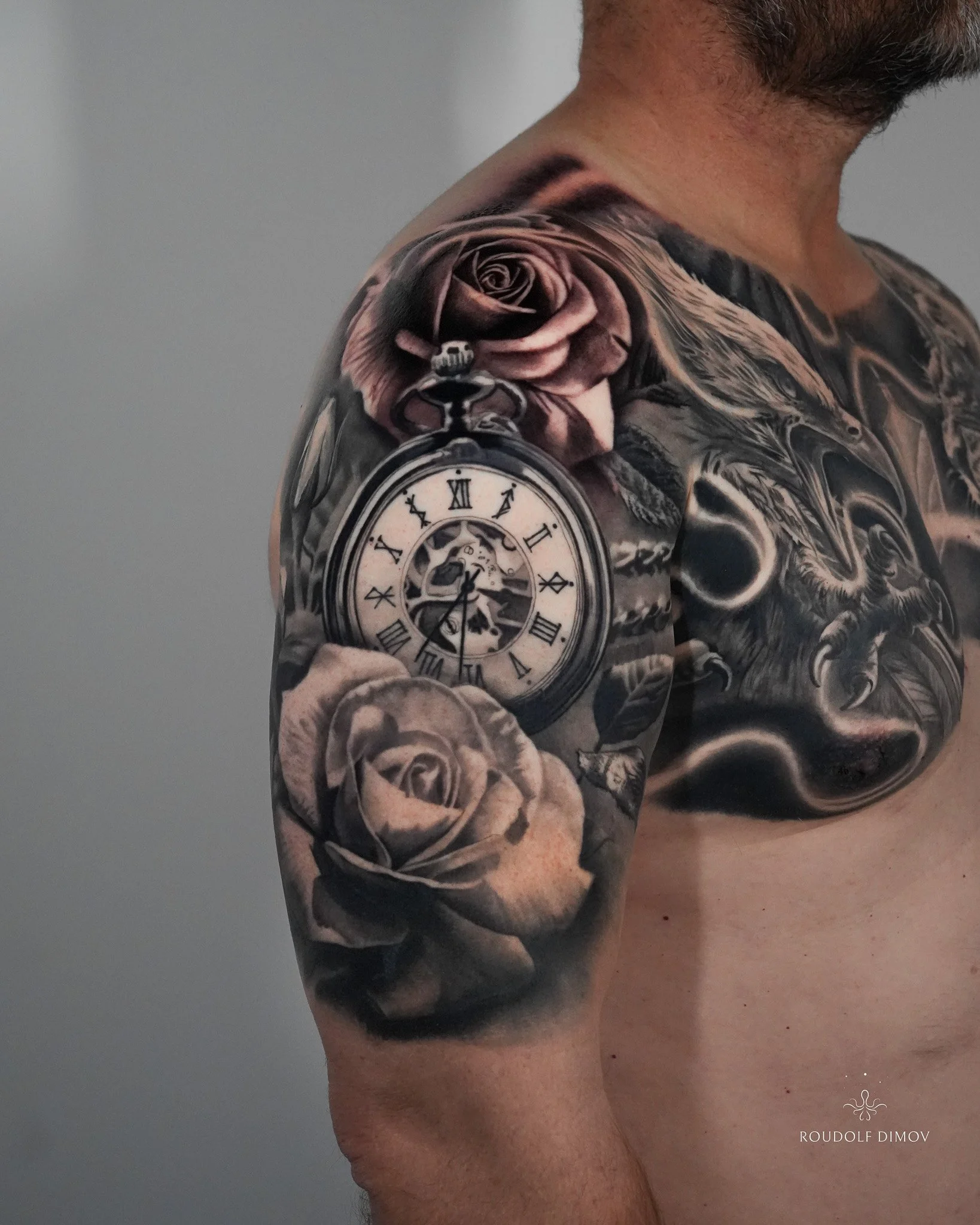 Black and grey realism upper-arm tattoo featuring a pocket watch, large roses, and subtle background flow connecting the design to the chest piece.