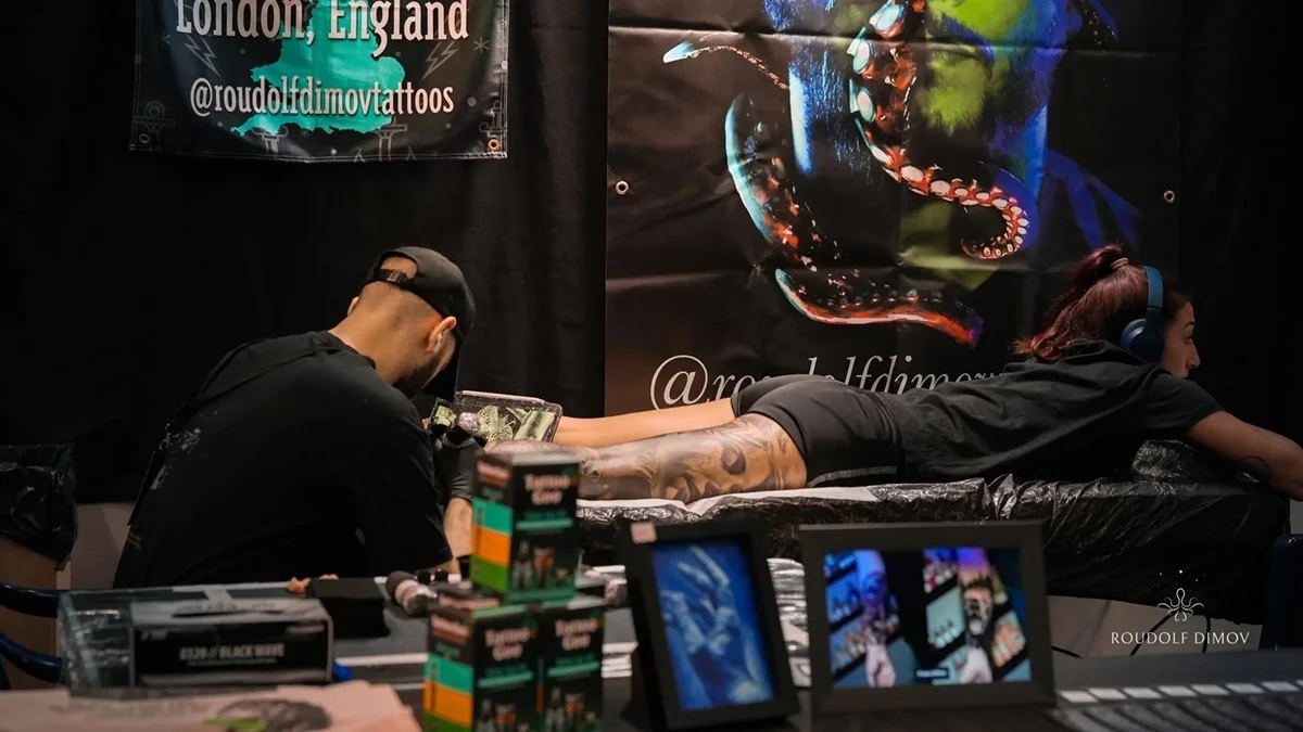 Tattoo artist working on a female client lying on a table with her arm extended. The artist is wearing a black cap and gloves, and the client is wearing headphones. Background banners display social media handles and artwork of an octopus tattoo.