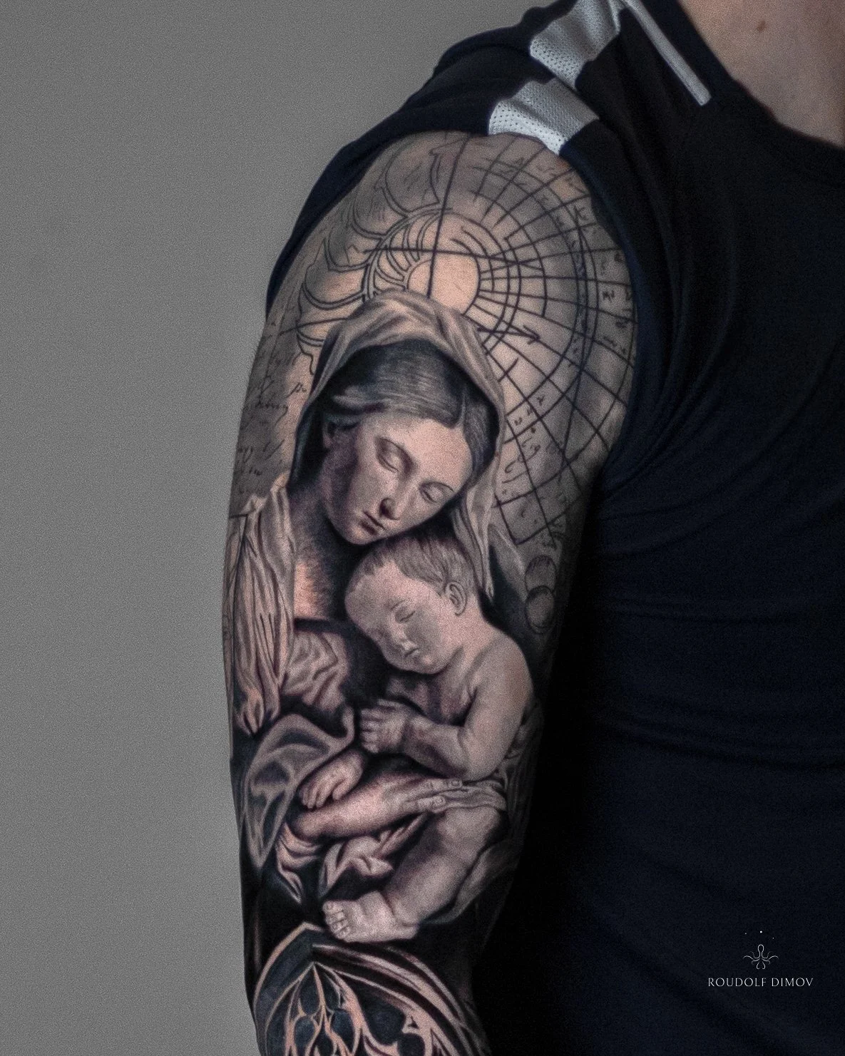 Black and grey realism tattoo of the Virgin Mary holding baby Jesus on the upper arm, framed with Renaissance-style sketchwork and soft shading as part of a full sleeve.
