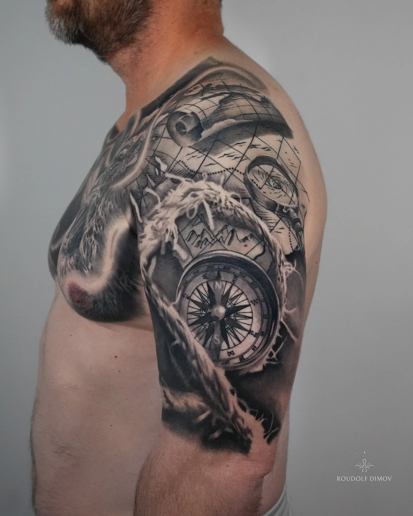 Black and grey realism upper-arm tattoo featuring a compass, vintage map, nautical rope, and magnifying glass, with negative-space flow connecting it to the chest piece.