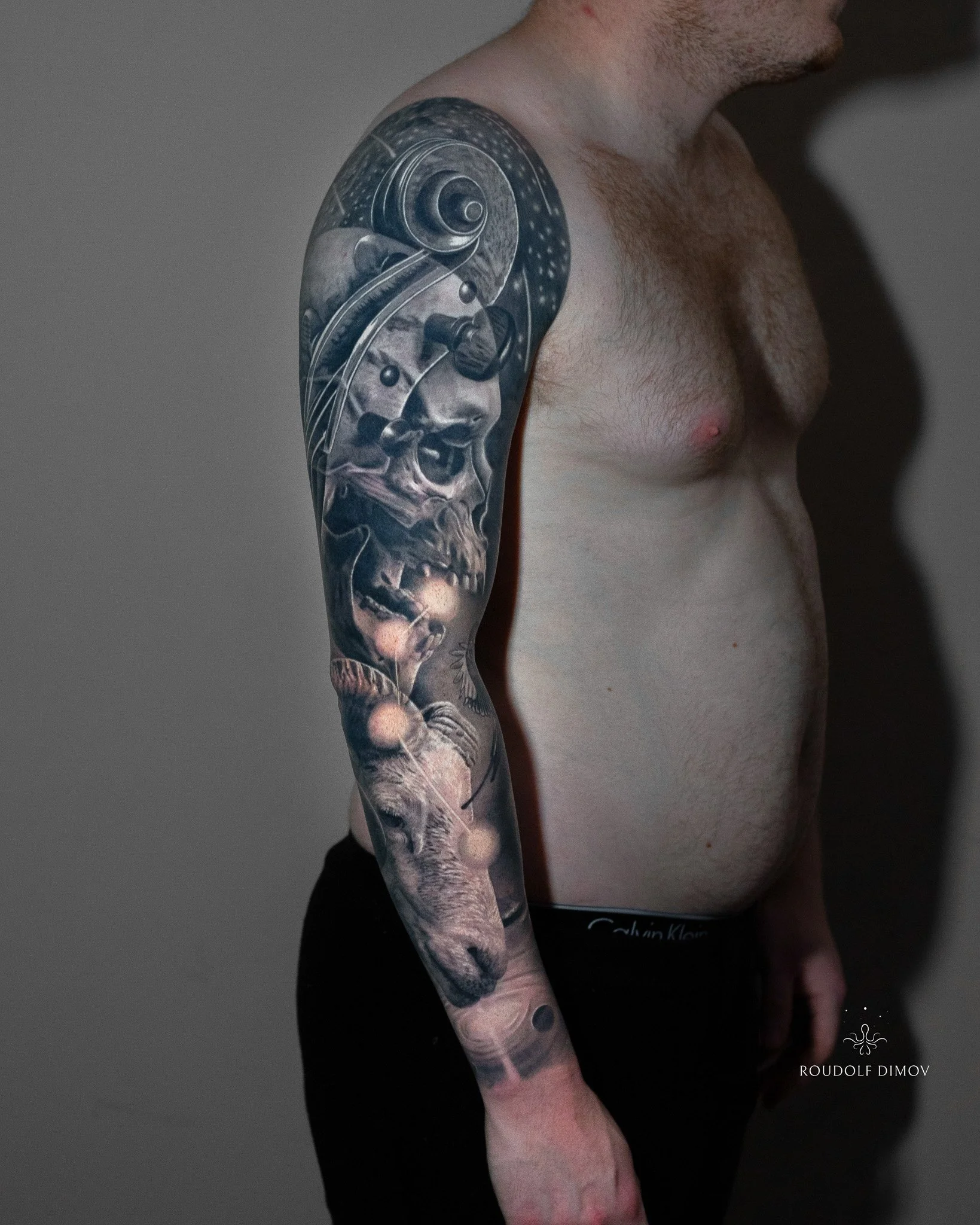 Black and grey realism full sleeve tattoo featuring a Capricorn goat, surreal skull with violin scroll, cosmic elements, and smooth transitions, created by London tattoo artist Roudolf Dimov.