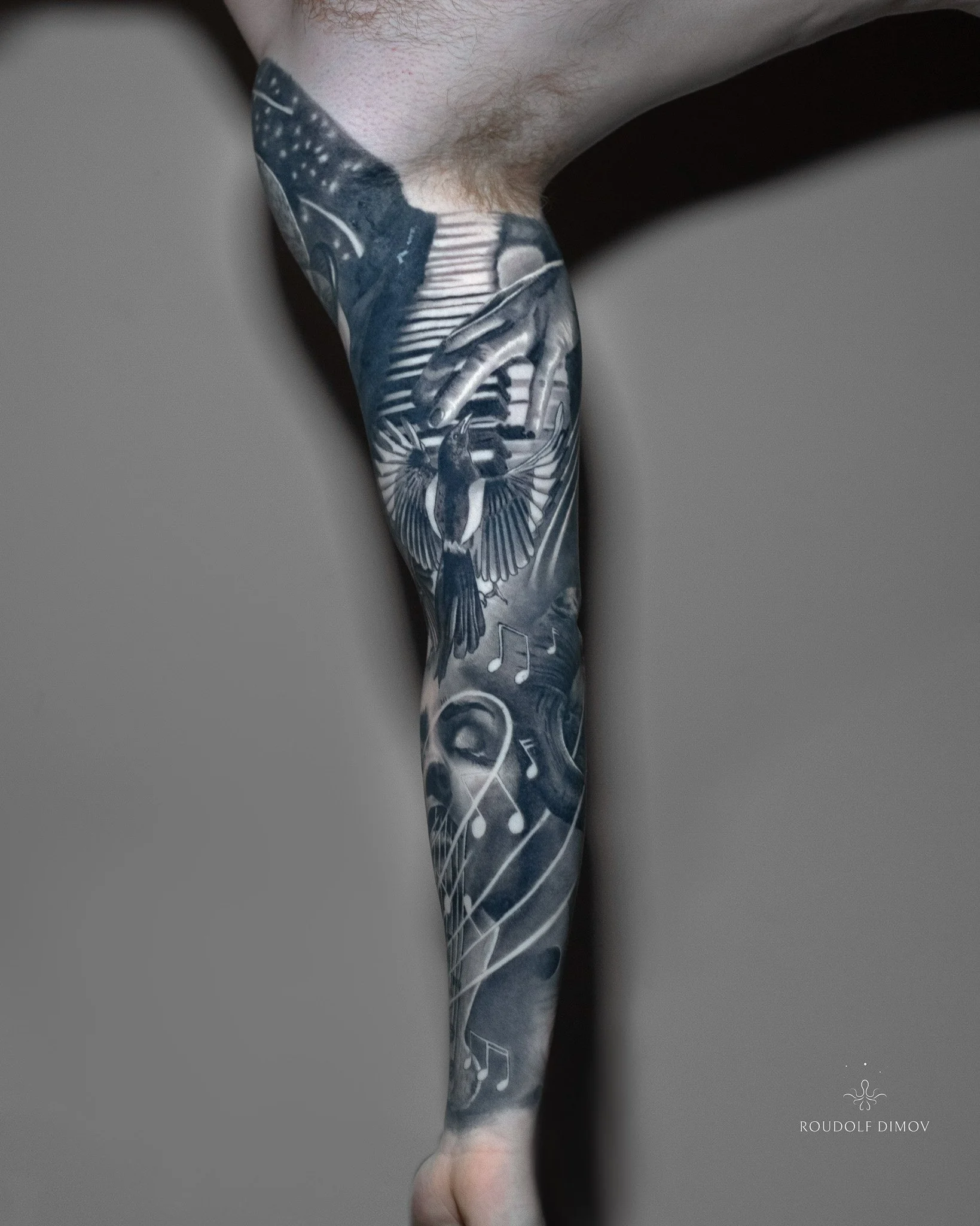 Black and grey realism inner arm tattoo featuring piano-playing hands, a flying magpie, musical notes, and flowing transitions, created by London tattoo artist Roudolf Dimov.