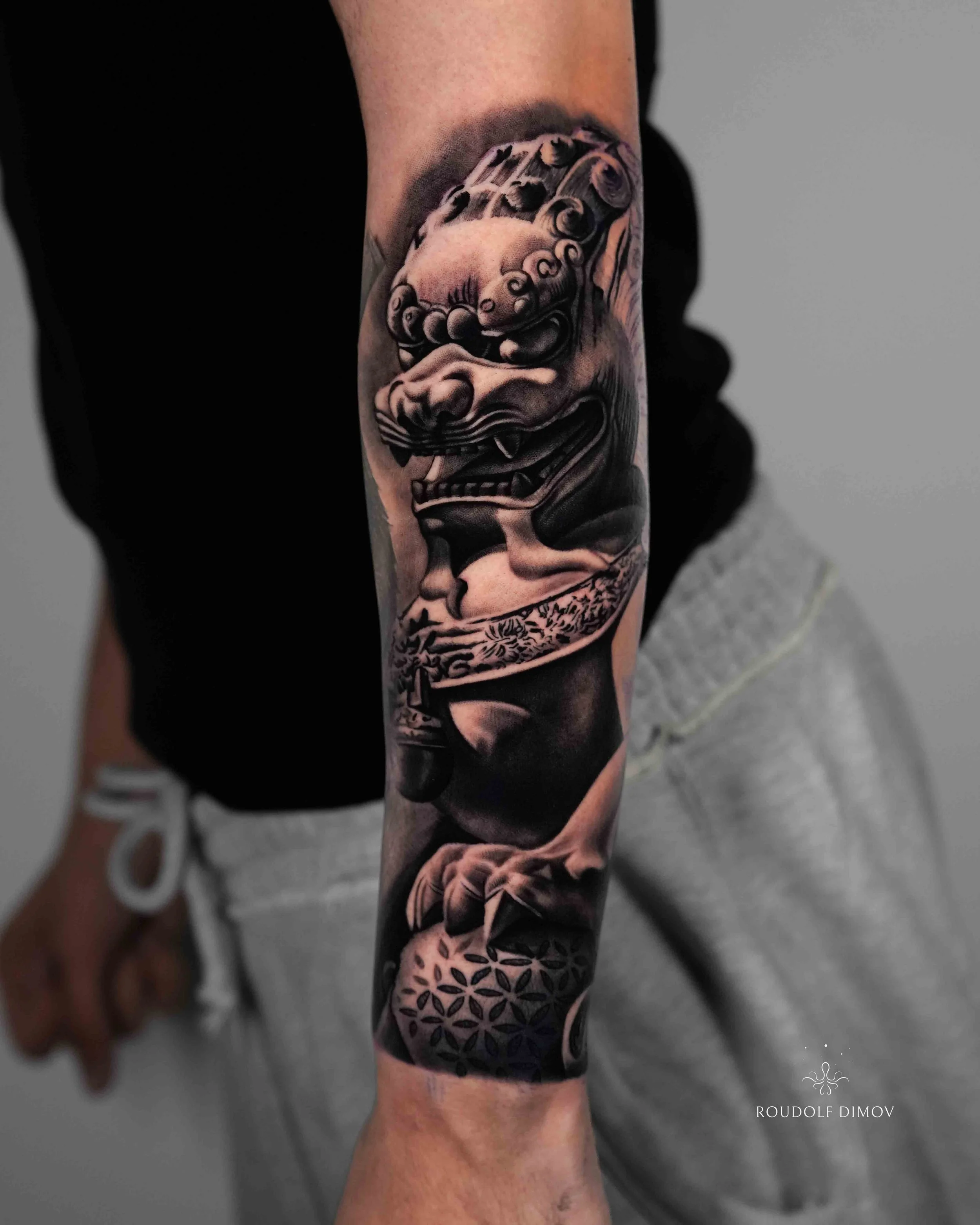 Black and grey realism Foo Dog tattoo on the forearm by London tattoo artist Roudolf Dimov. This Japanese guardian lion design combines traditional symbolism with detailed realism, featuring smooth shading, high contrast, and bold textures. The tatto