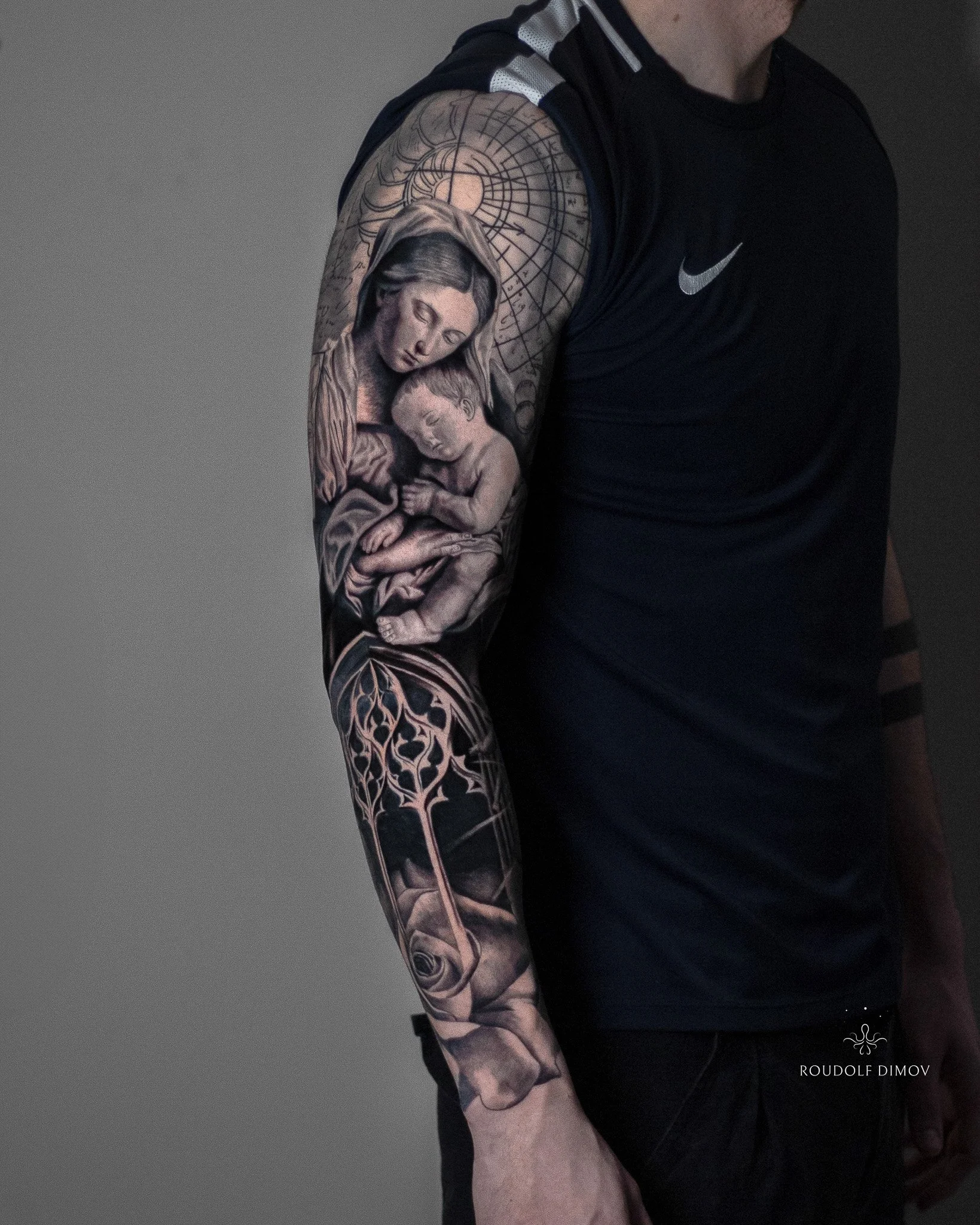 Black and grey realism tattoo of the Virgin Mary with baby Jesus on the upper arm, flowing into a rose window and rose on the forearm as part of a full sleeve.