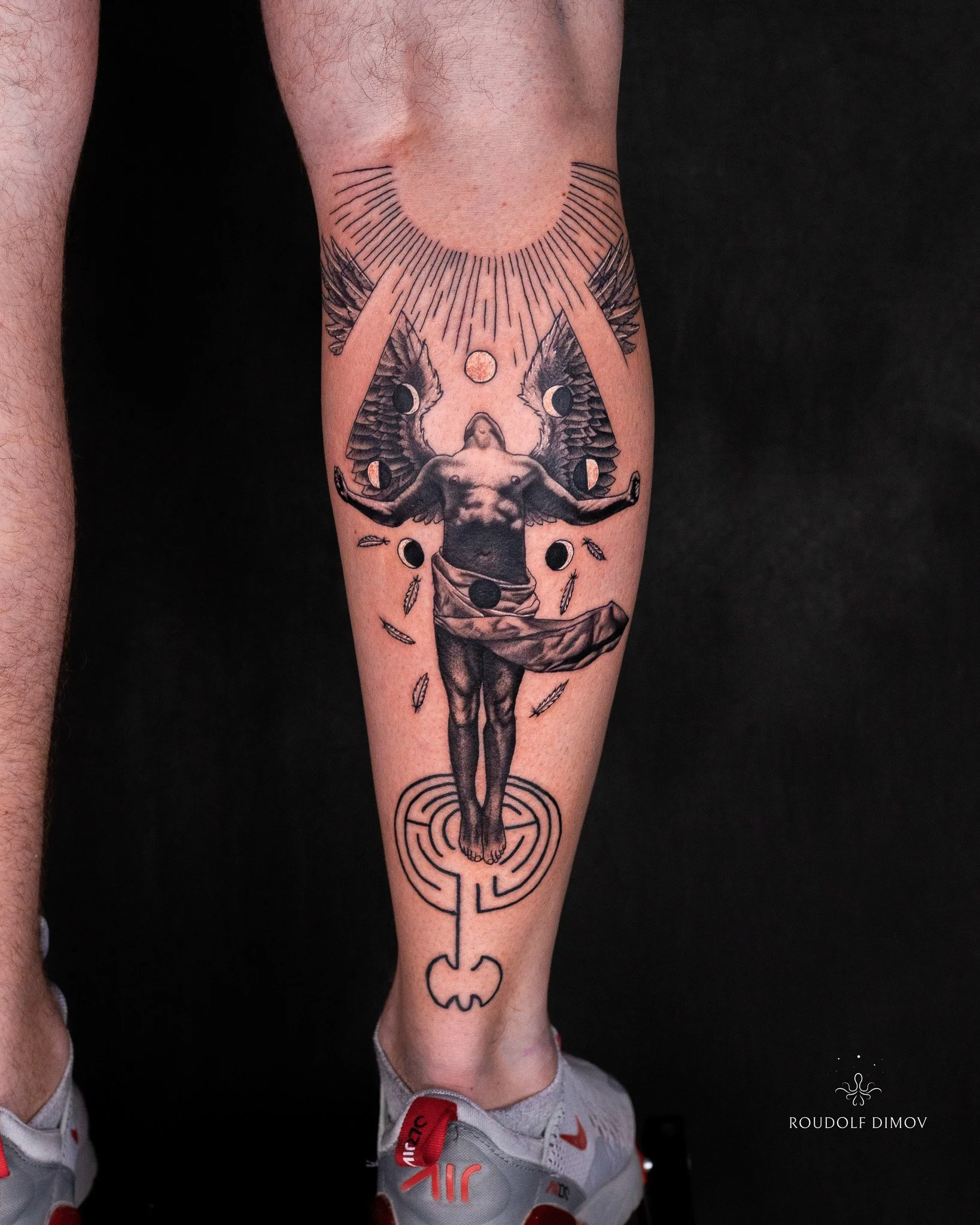 Black and grey realism calf tattoo of Icarus rising toward the sun, featuring stylised wings, falling feathers, lunar phases, and a labyrinth symbol beneath his feet.