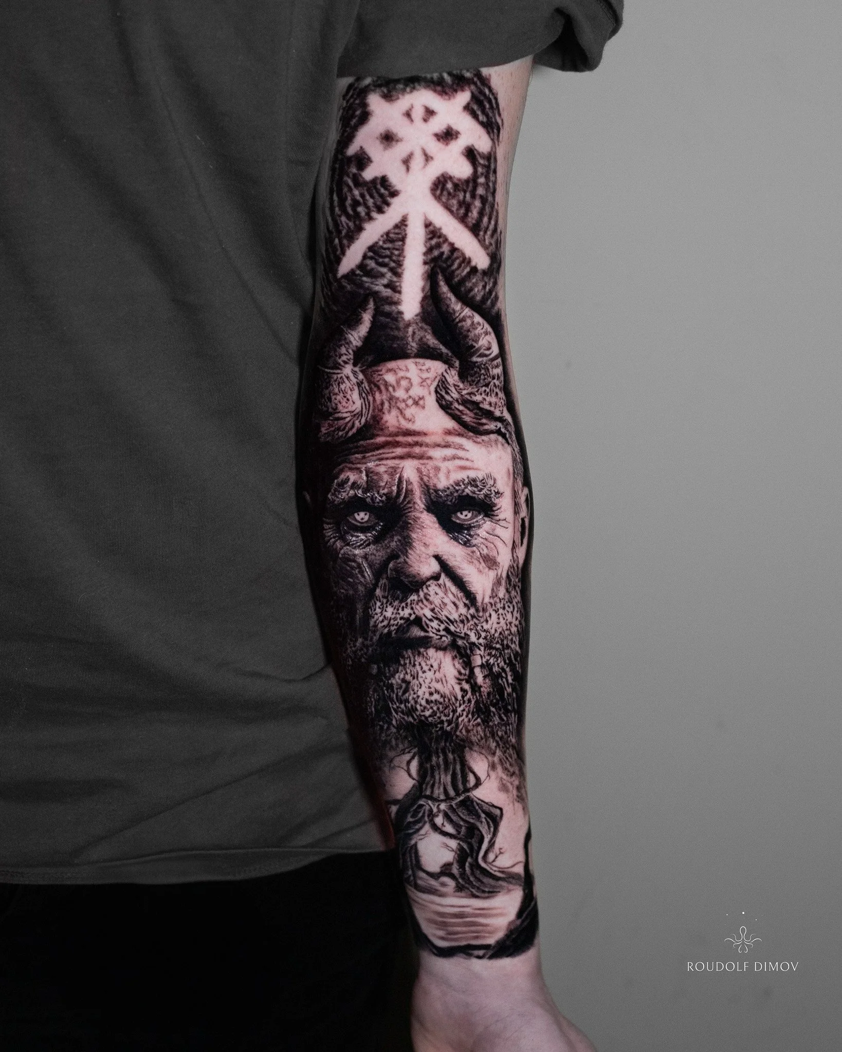 Mimir tattoo on the forearm in black and grey realism, featuring Norse mythology symbolism, detailed portrait work, and runic elements. Tattoo artist Roudolf Dimov, London.