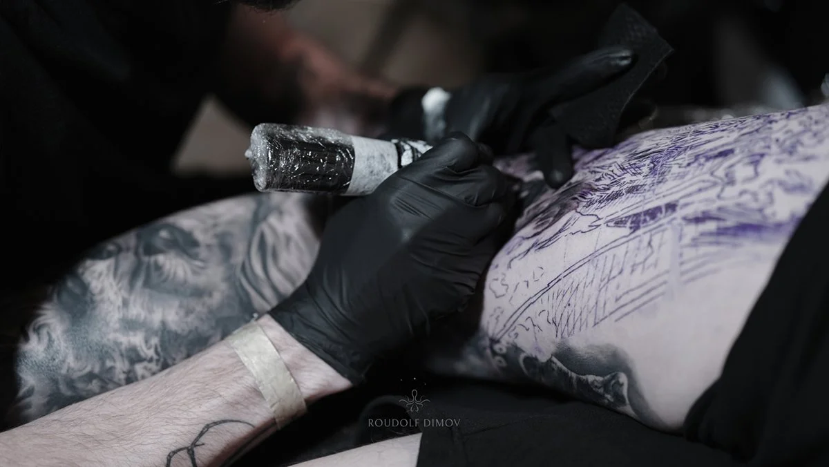 A tattoo artist wearing black gloves is creating a tattoo on a person's side torso using a tattoo machine. The person's skin is stretched, and the tattoo design appears to be detailed and intricate.