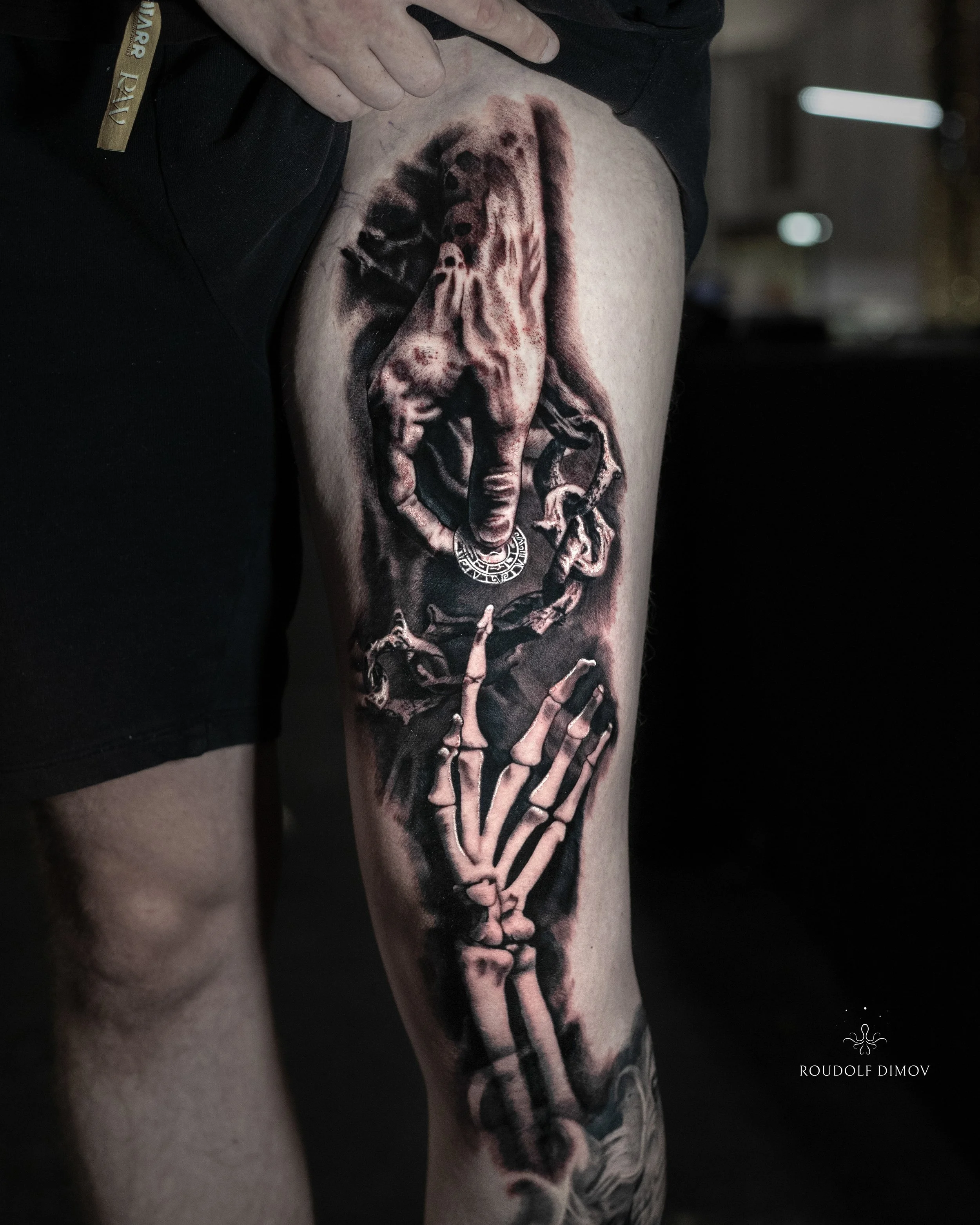 Skeleton hand and mythology symbol tattoo on the thigh in black and grey realism, showing detailed bone structure and dramatic shading. Tattoo artist Roudolf Dimov, London.