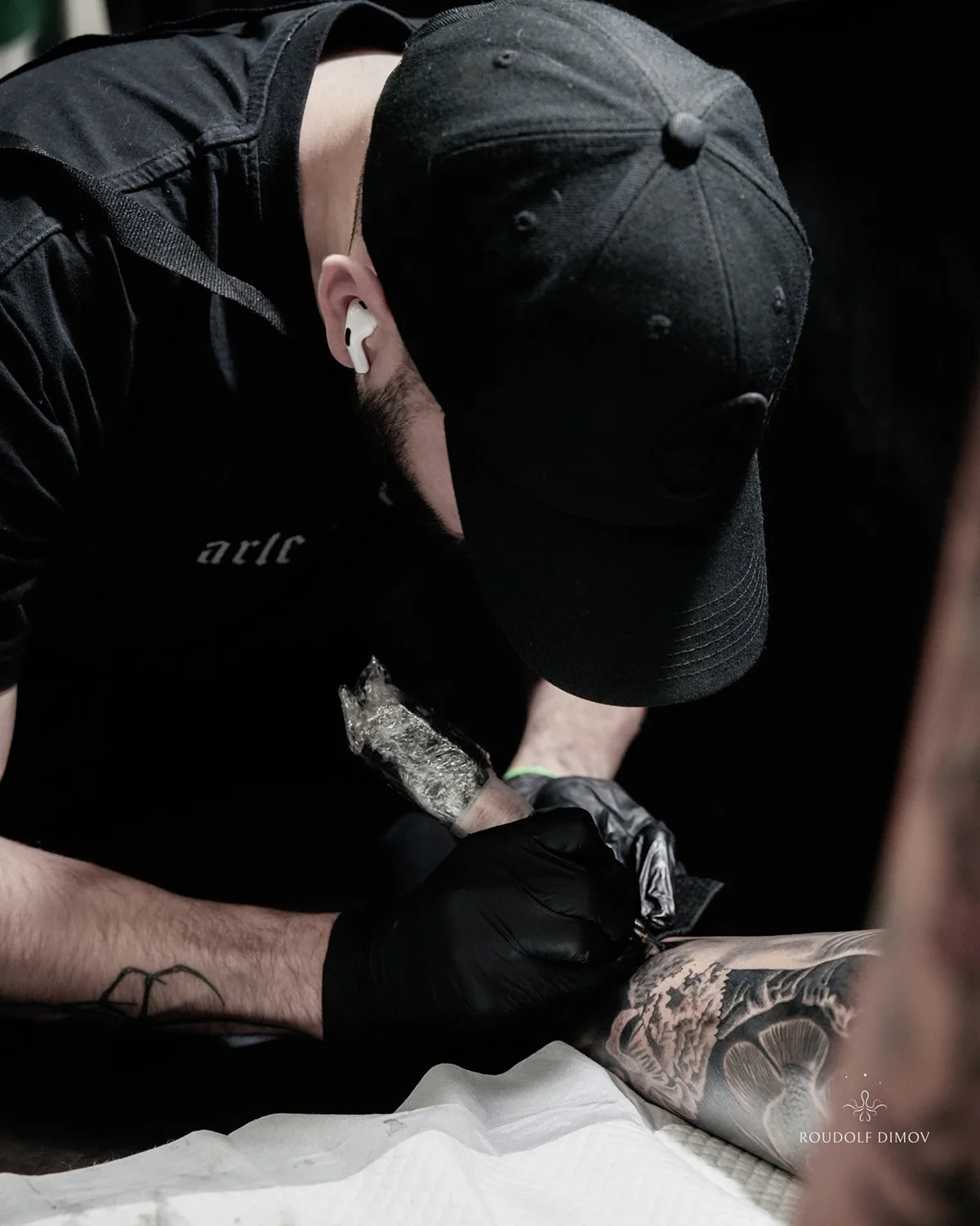 A tattoo artist is creating a detailed black and gray sleeve tattoo on a person's arm using a tattoo machine. The artist is wearing black gloves and a black cap, and has a beard. The tattoo features intricate shading and realistic elements.
