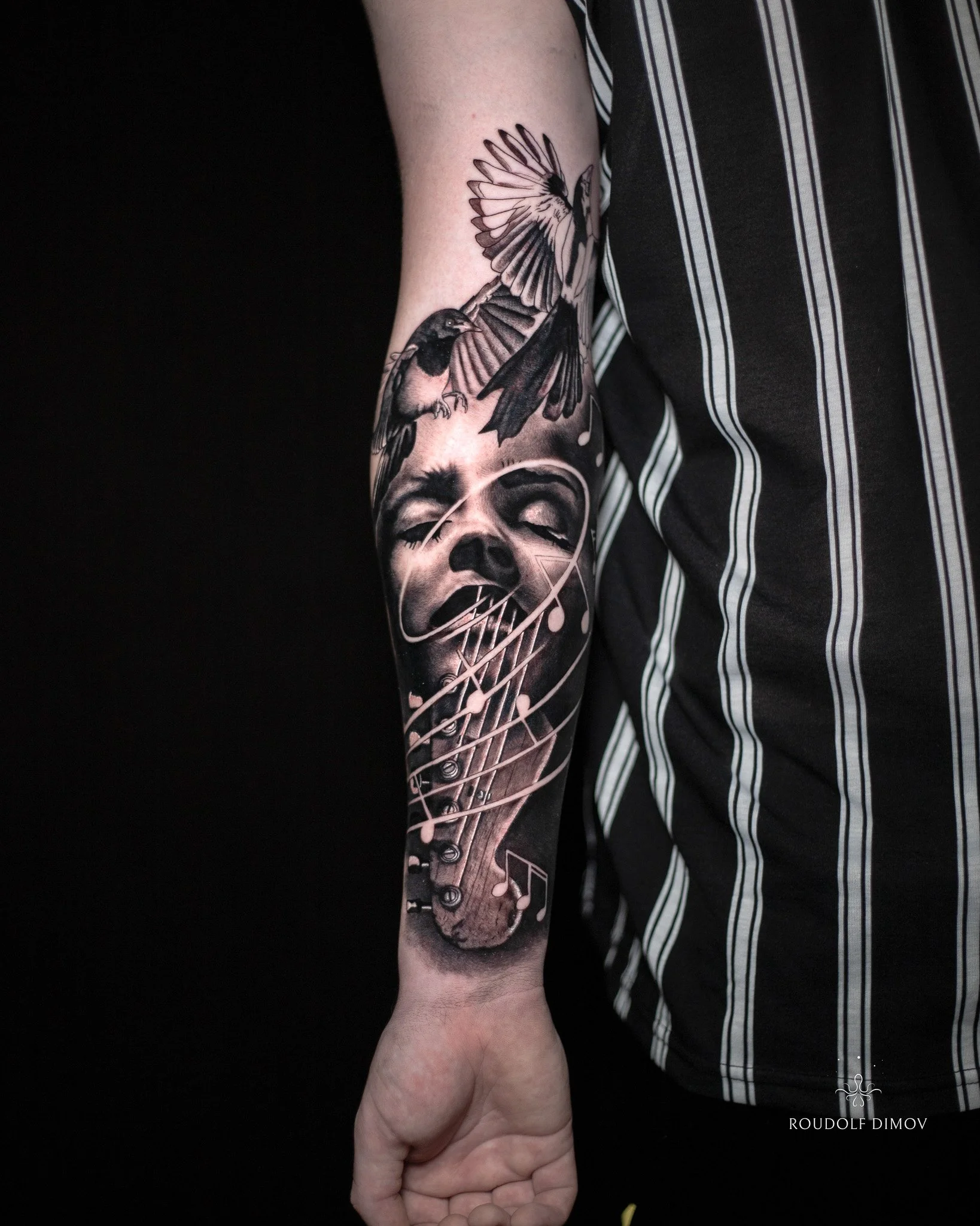 Black and grey realism forearm tattoo featuring two magpies, a female face, musical notes, and a left-handed guitar, created by London tattoo artist Roudolf Dimov.