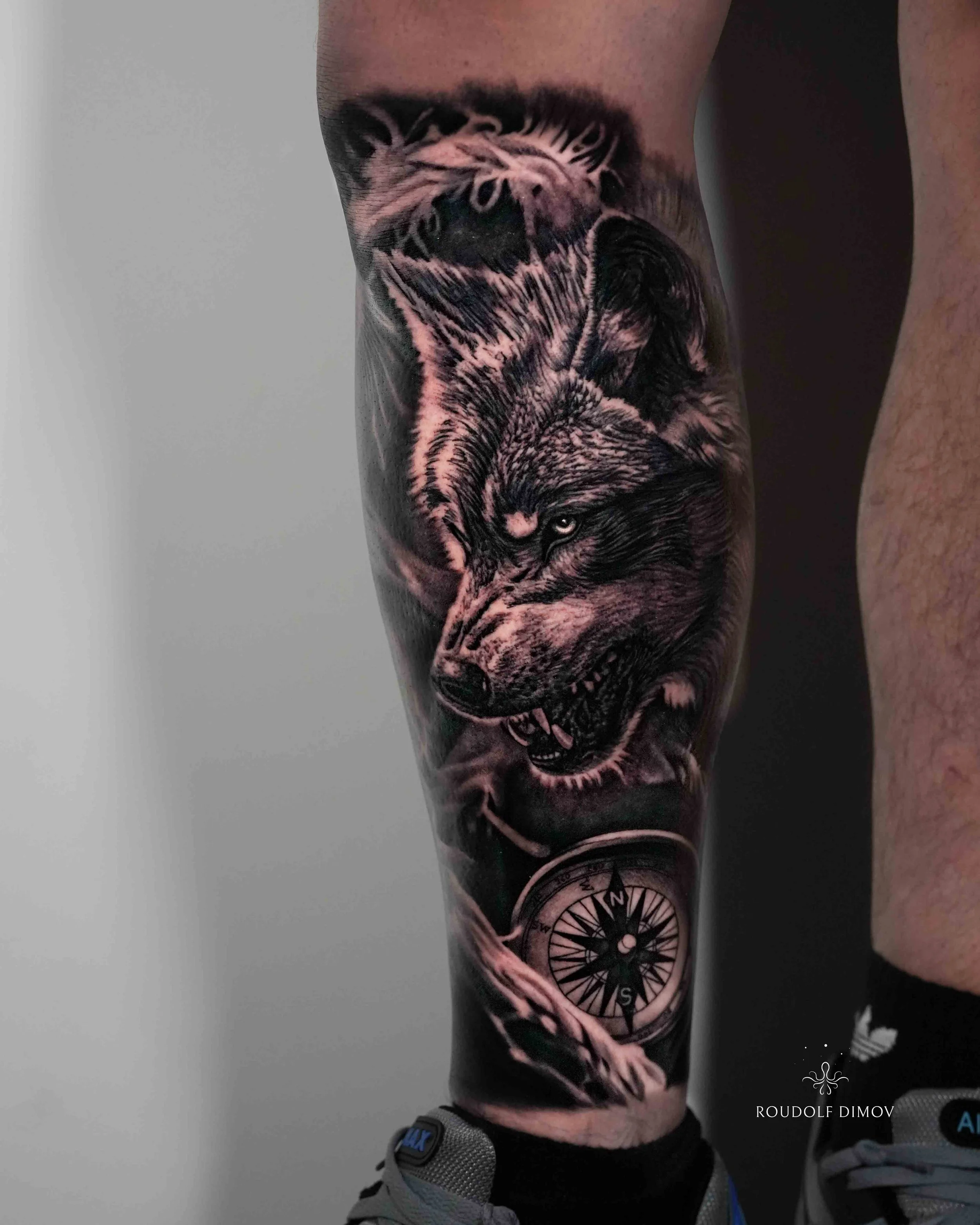 Black and grey realism wolf tattoo on the calf featuring a detailed compass design by London tattoo artist Roudolf Dimov. This custom leg tattoo combines the raw power of a snarling wolf with the symbolism of guidance, instinct, loyalty, and survival
