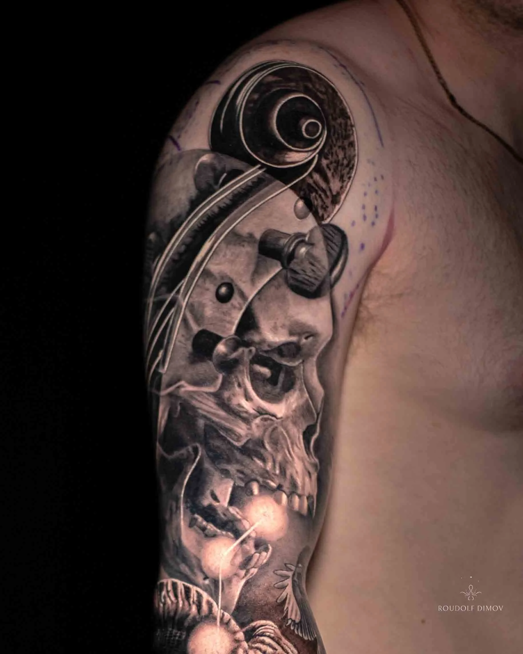Black and grey realism upper arm tattoo featuring a surreal skull blended with a violin scroll and cosmic elements, created by London tattoo artist Roudolf Dimov.