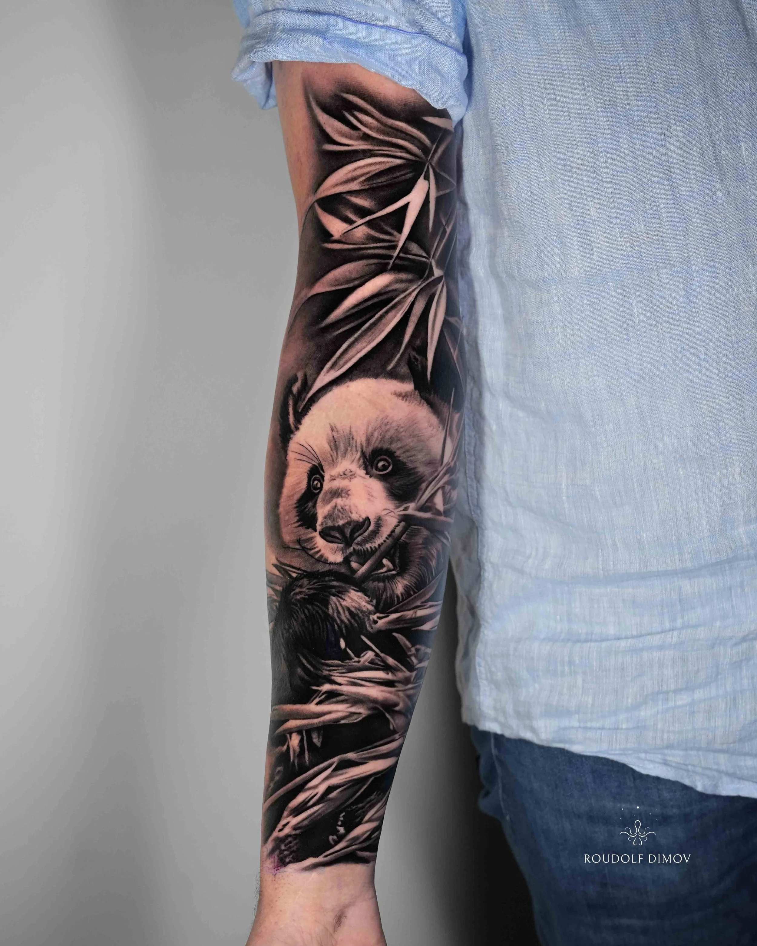 Black and grey realism panda tattoo sleeve on the forearm by London tattoo artist Roudolf Dimov. This detailed panda tattoo is surrounded by flowing bamboo leaves, creating a natural Japanese-inspired composition with strong contrast and smooth shadi