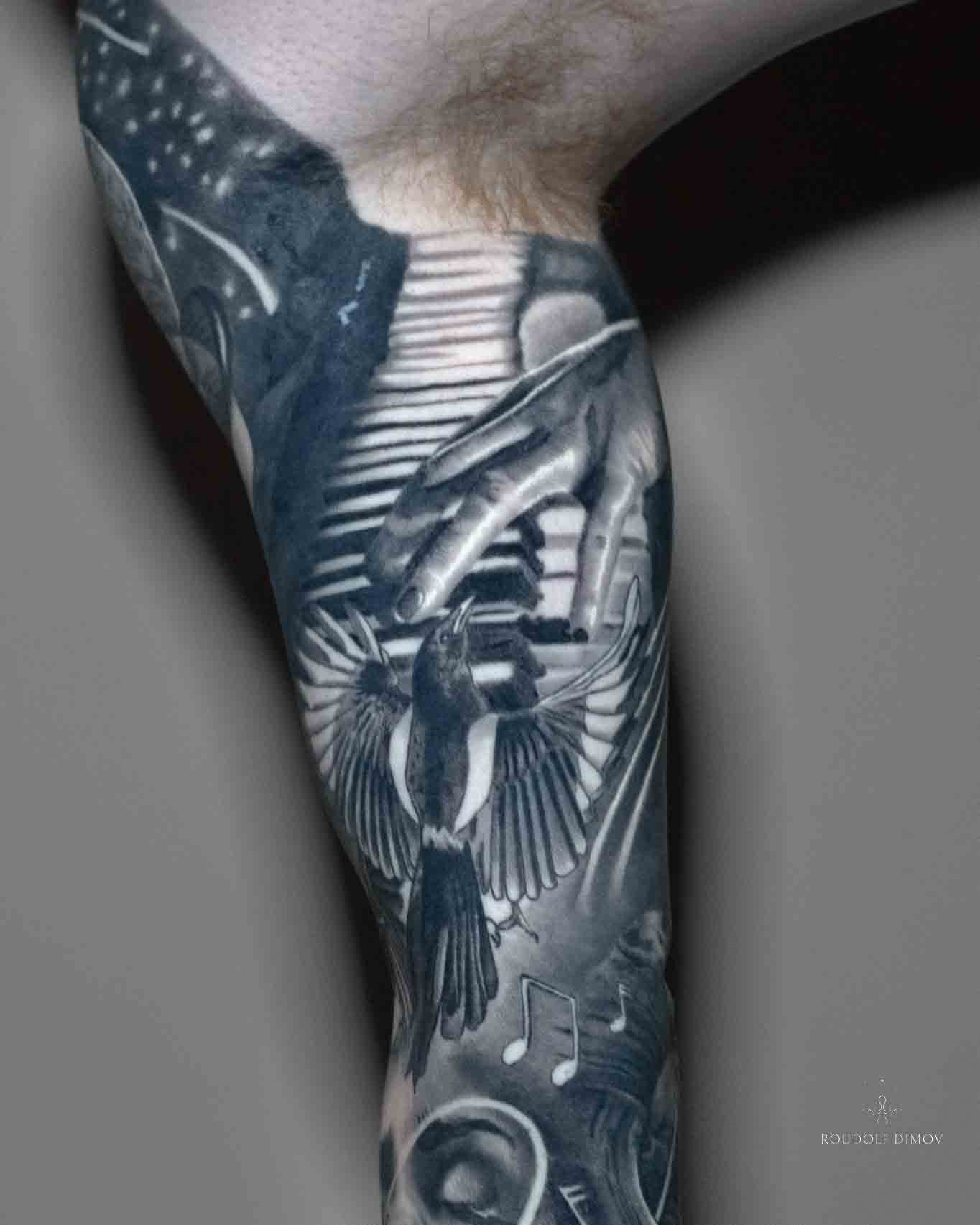 Black and grey realism inner arm tattoo featuring piano-playing hands, a magpie, musical notes, and cosmic transitions, created by London tattoo artist Roudolf Dimov.