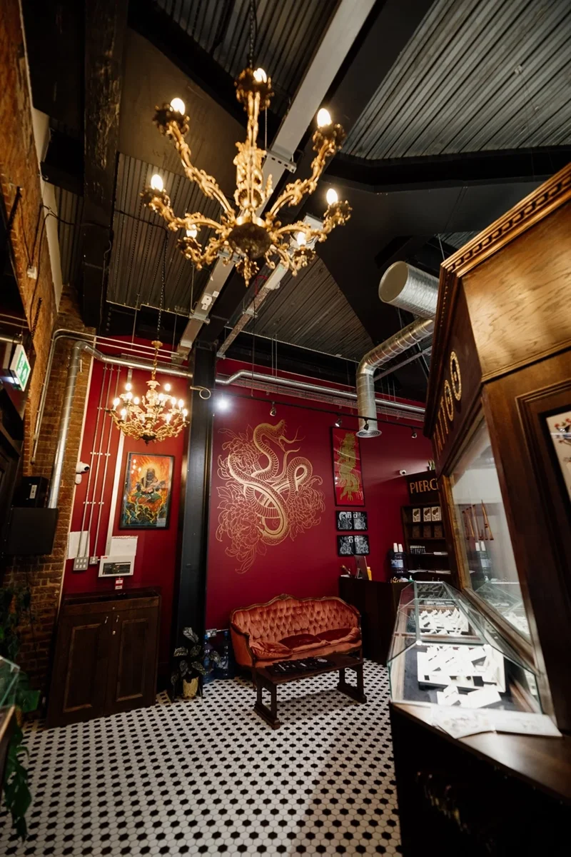 Interior of a restaurant or cafe with black and white checkered tile floor, dark ceiling, red walls decorated with dragon artwork, vintage chandelier, plush pink velvet sofa, and display case.