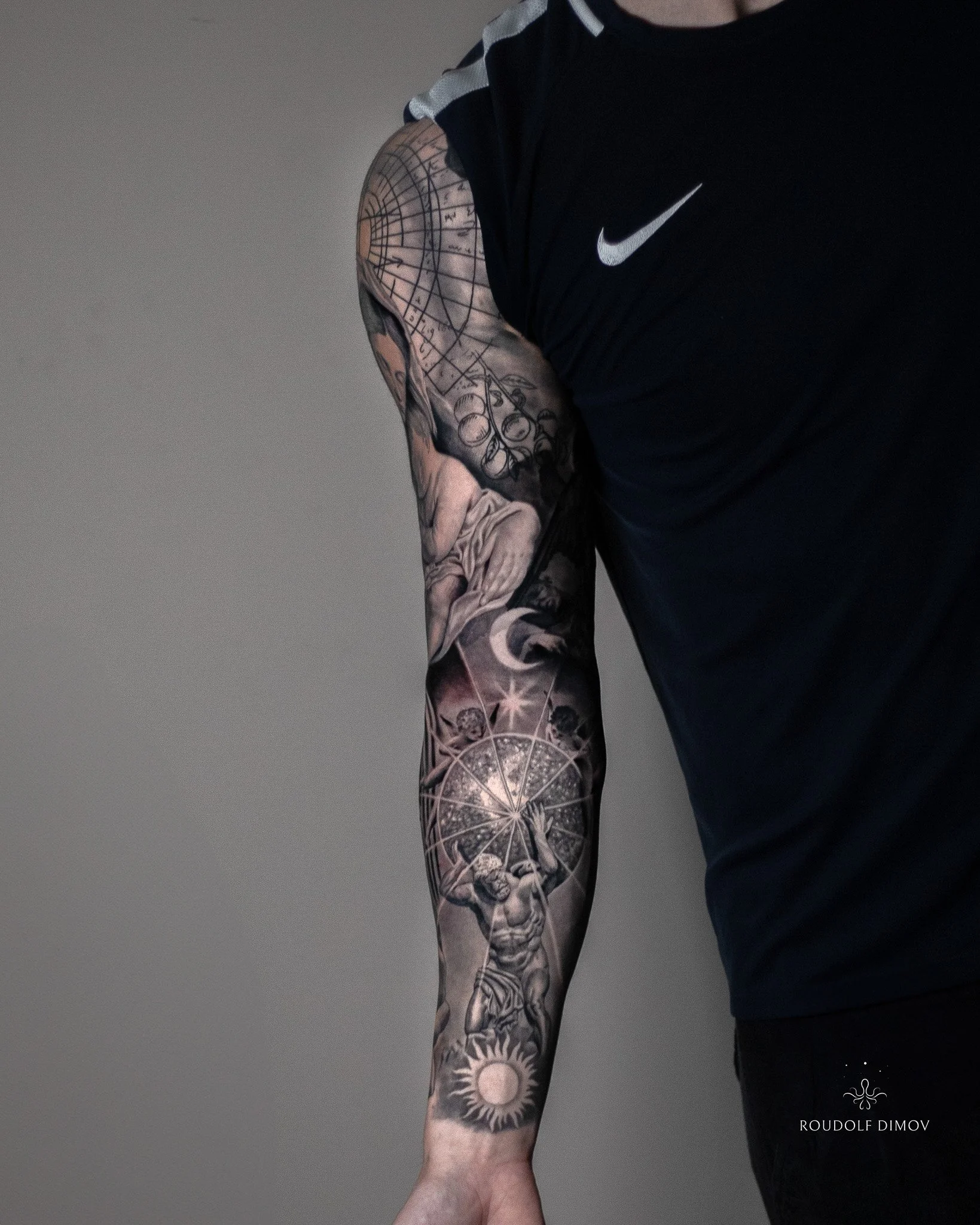 Full black and grey realism sleeve showing Atlas with a celestial sphere, angels, the moon and sun, flowing into Renaissance elements on the upper arm.