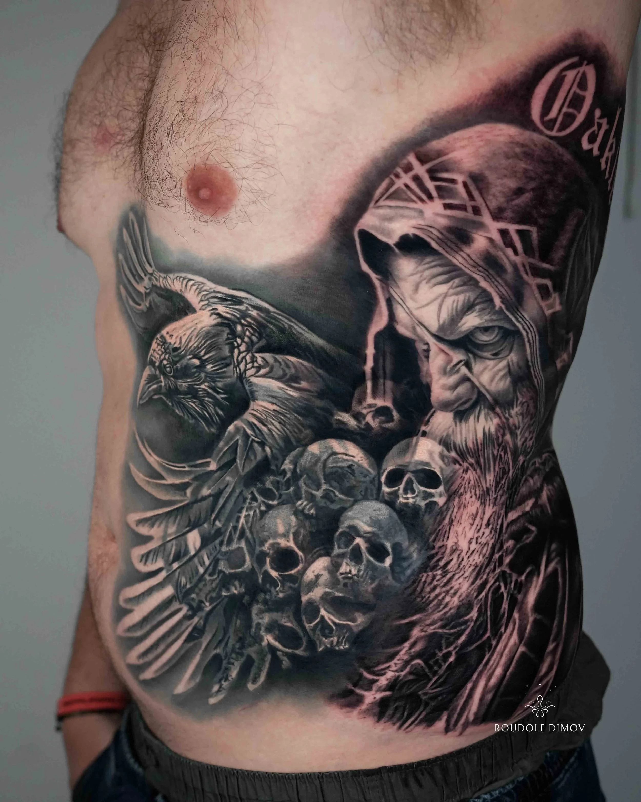 Black and grey realism Norse mythology rib tattoo featuring Odin, a raven, and detailed skull composition by London tattoo artist Roudolf Dimov. This dark atmospheric tattoo combines Viking symbolism with realistic texture, strong contrast, and smoot