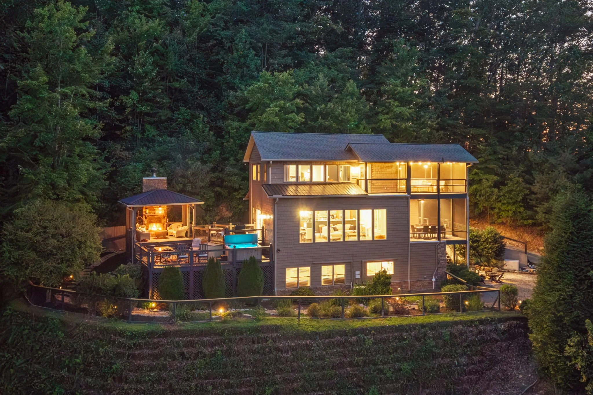 A multi-story house with large windows and a balcony, illuminated from within, located in a wooded area at dusk. The house has a patio with outdoor furniture and a backyard with a swimming pool.
