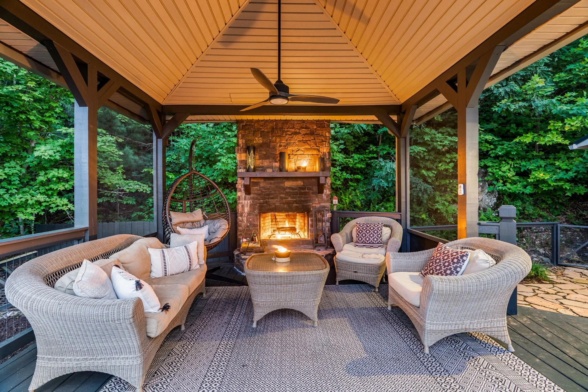 Outdoor covered patio with wicker furniture, a brick fireplace, and green trees in the background. There's a ceiling fan and warm lighting.