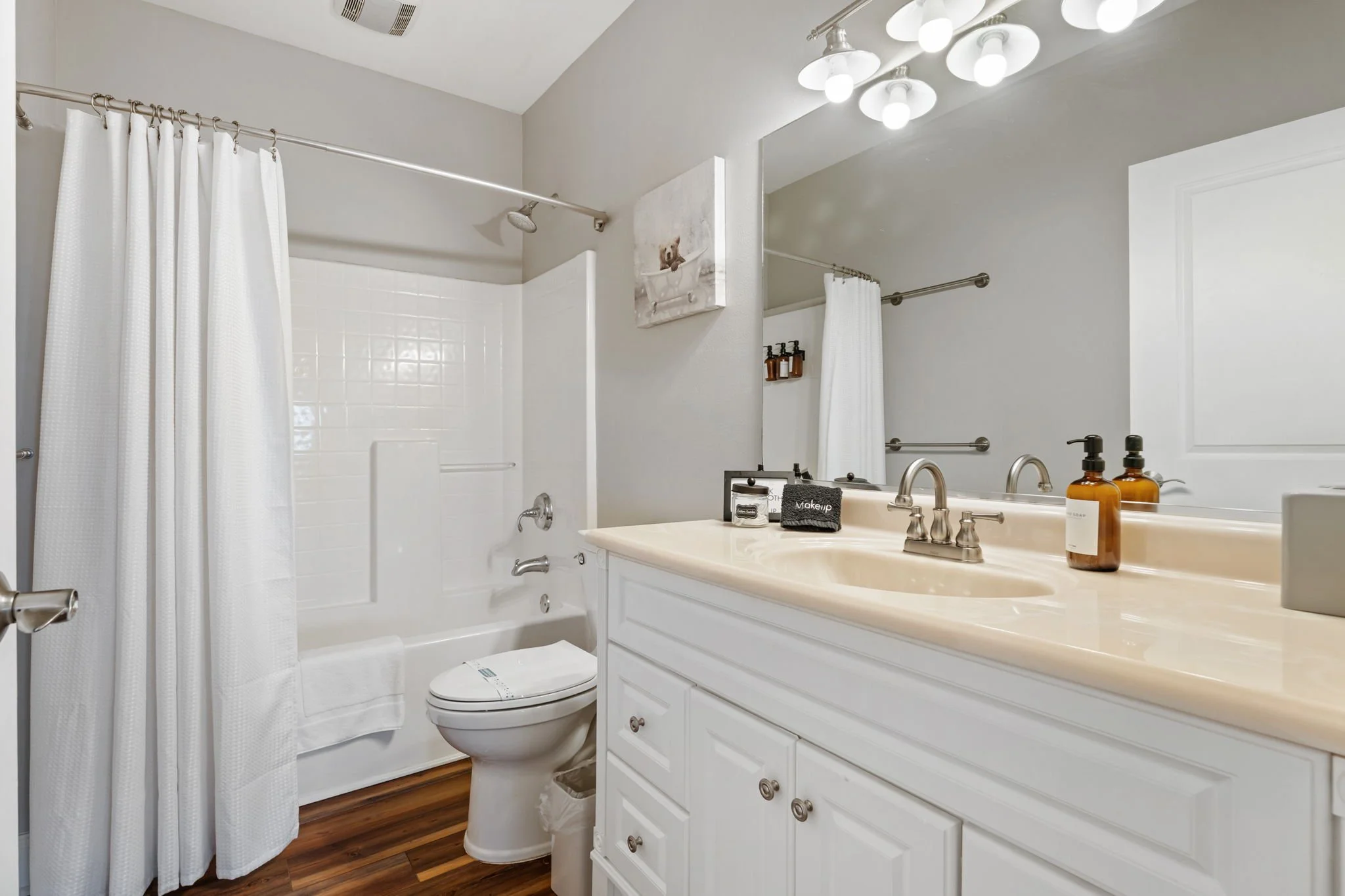 Bathroom with white vanity, soap and lotion bottles, mirror with light fixtures, toilet, bathtub with white shower curtain, and wall art with a kitten in a bathtub
