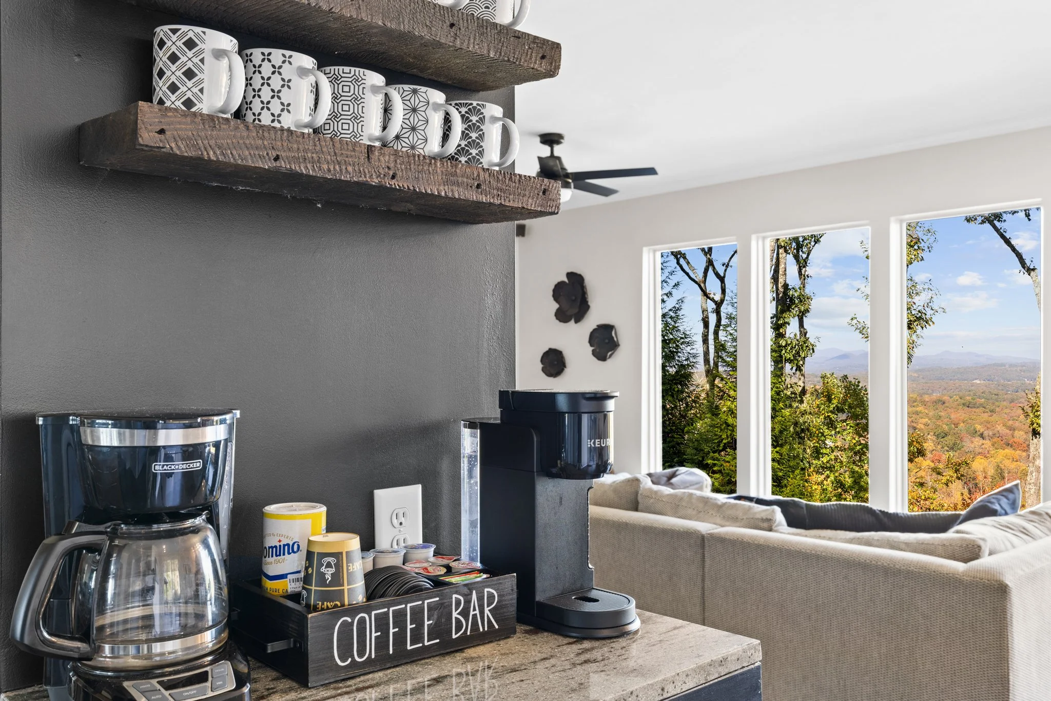 Kitchen countertop with coffee maker, coffee supplies, and a sign reading 'Coffee Bar.' Wall-mounted wooden shelves holding patterned coffee mugs. Large windows reveal trees and a scenic landscape outside.
