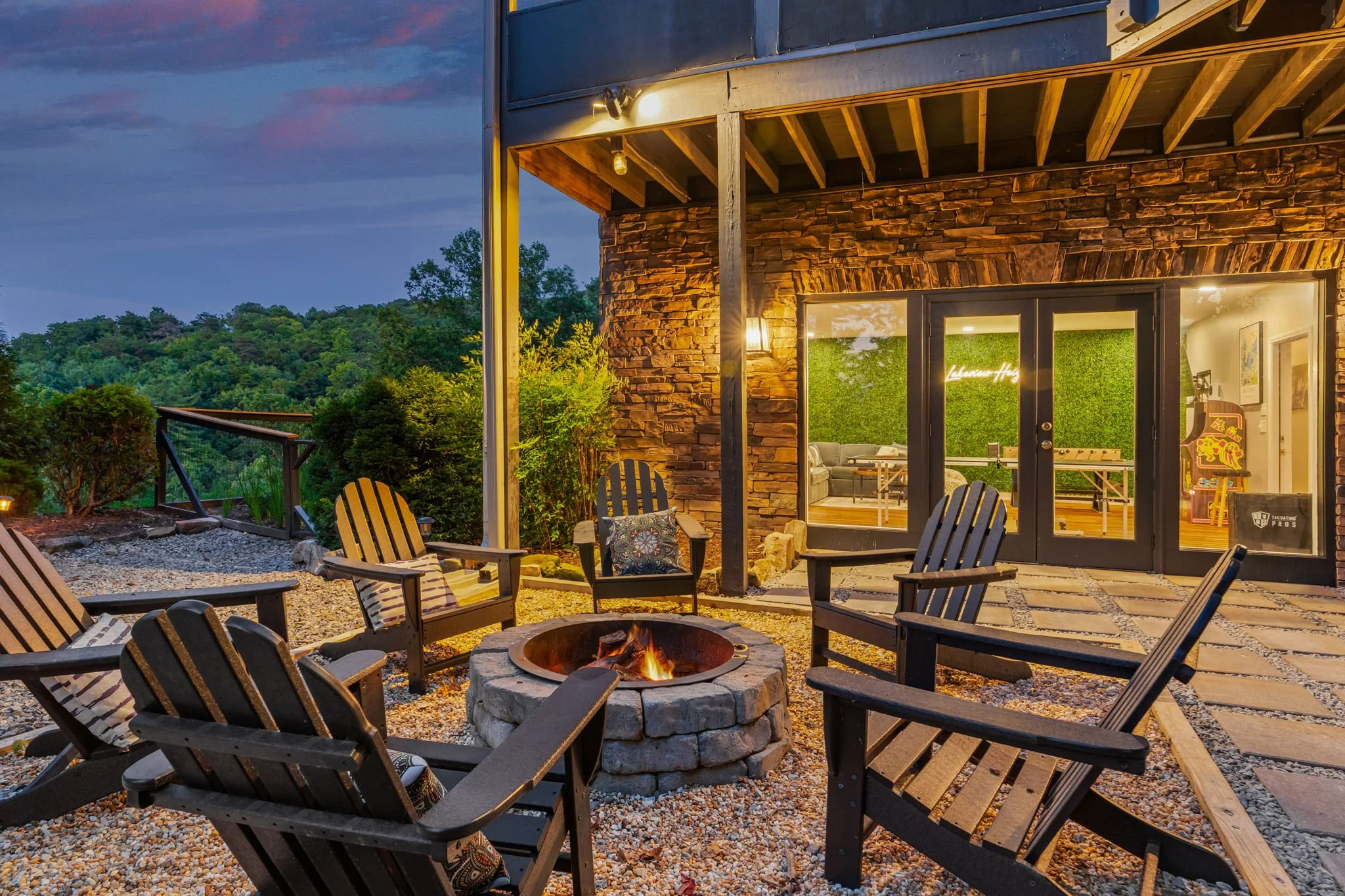 An outdoor patio area with a fire pit surrounded by wooden chairs. There is a house with a stone exterior and glass doors revealing a cozy interior with green wall decor. The scene is lit by warm lighting at dusk, with trees and a hill visible in the background.