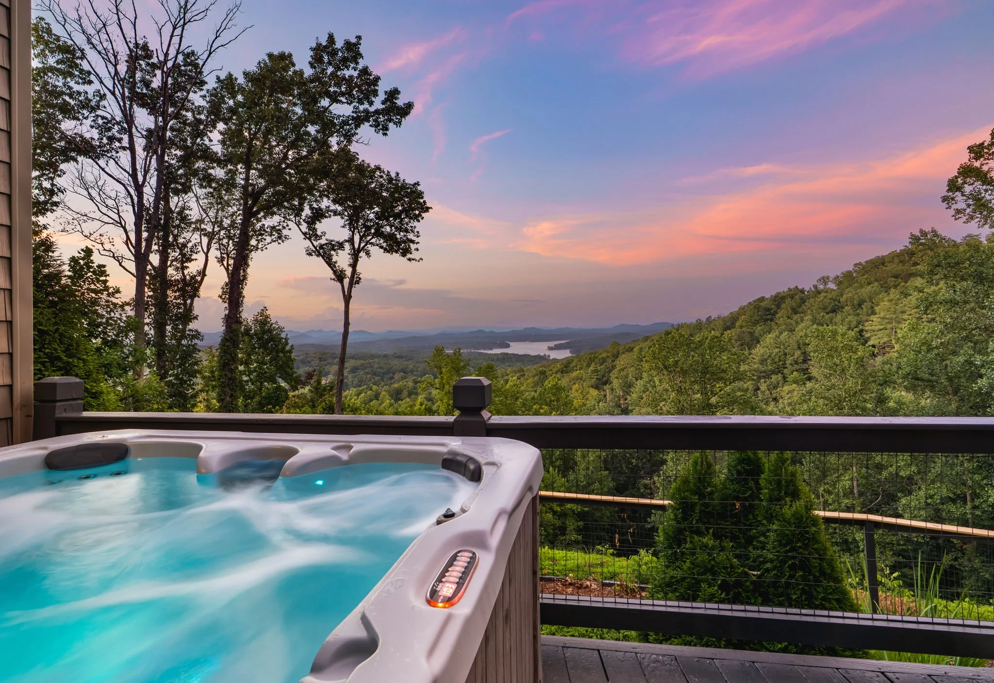 A hot tub on a wooden deck overlooking a scenic landscape with rolling hills, a river, and a colorful sunset sky with pink, purple, and blue clouds.
