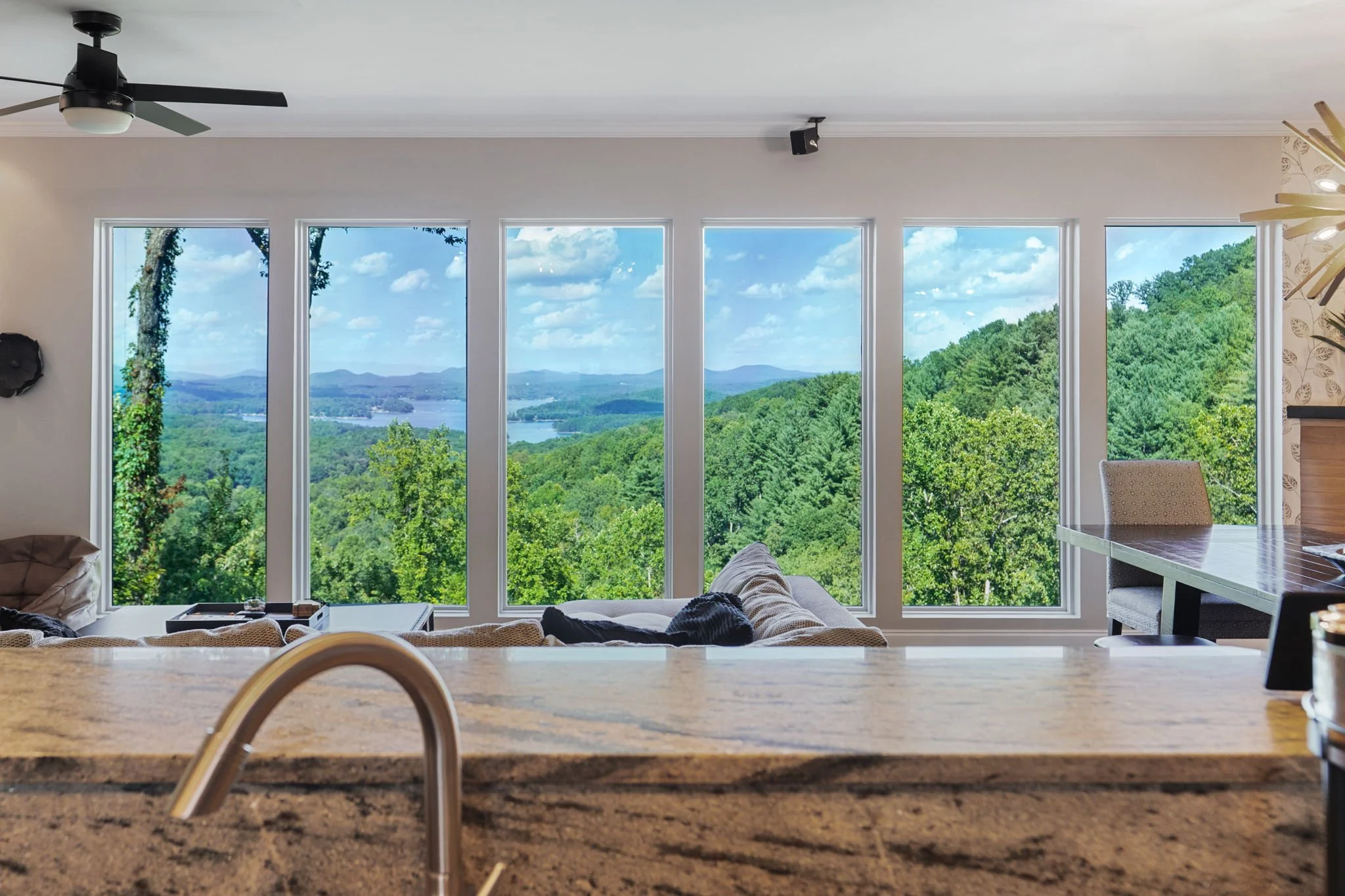 Living room with large windows showing a scenic view of green trees, hills, and a lake, with partial furniture including a sofa, a table, and a dining area.