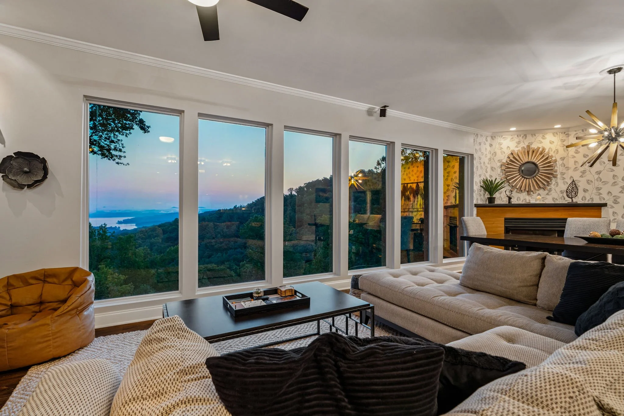 Living room scene with large windows showcasing a scenic view of trees and water during sunset, featuring modern furniture, a ceiling fan, decorative wall art, and plants.