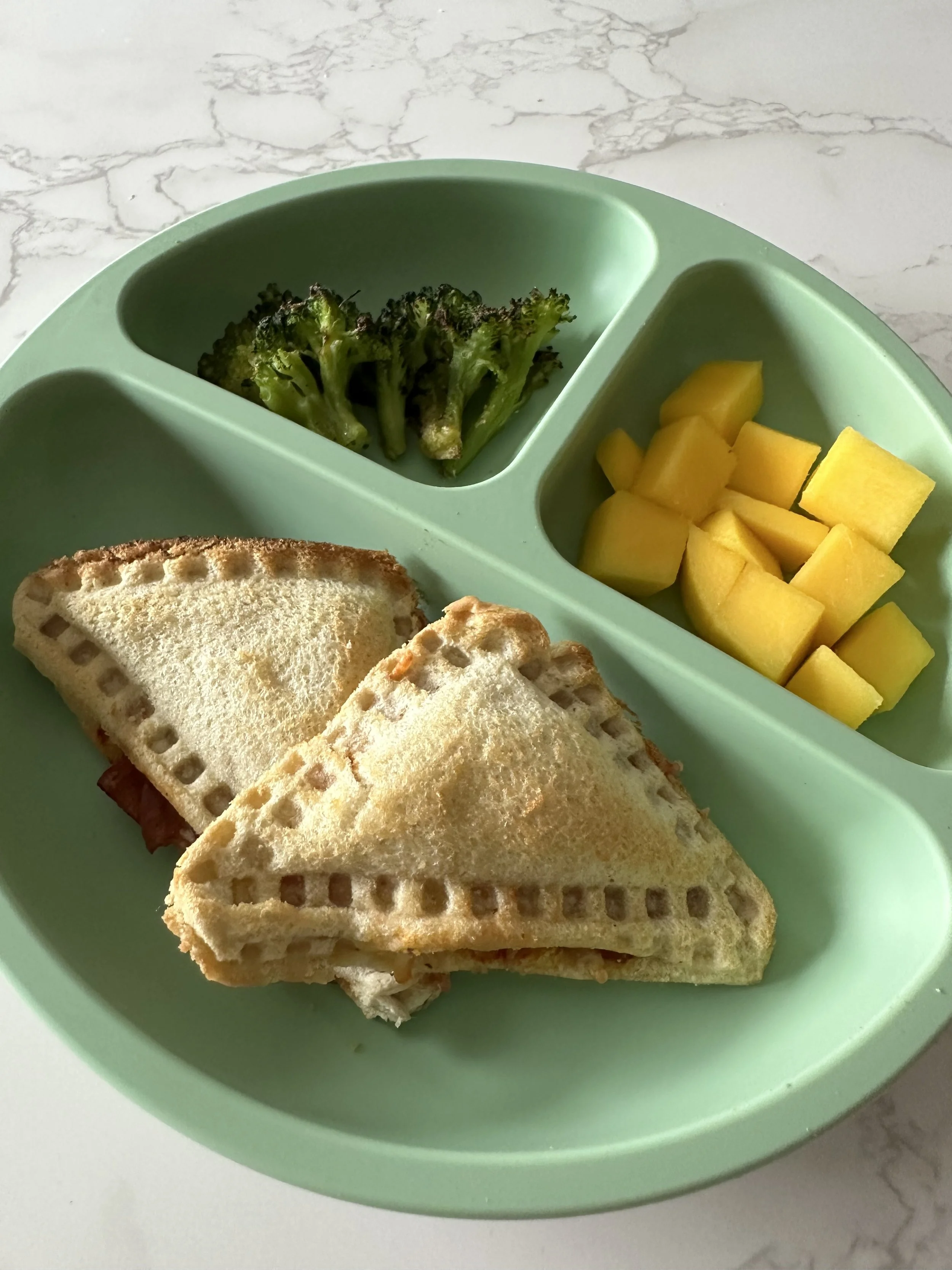 Green silicone suction plate with two air fried pizza uncrustable triangles, roasted broccoli, and fresh mango.