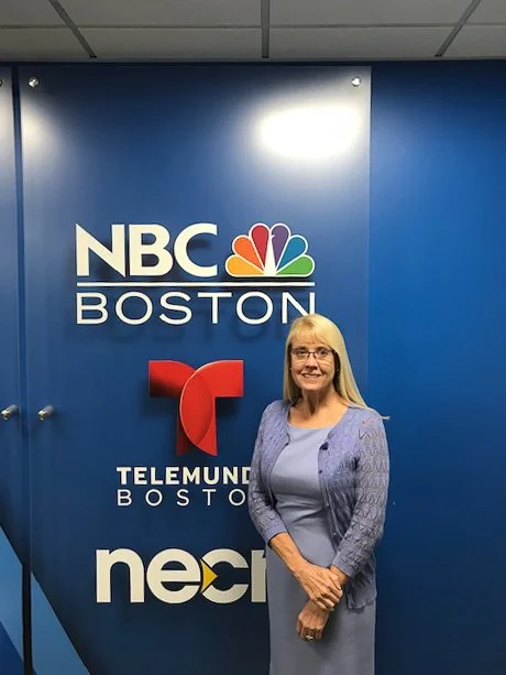 A woman with blonde hair and glasses standing in front of a blue wall with NBC Boston, Telemundo Boston, and NEC logos.