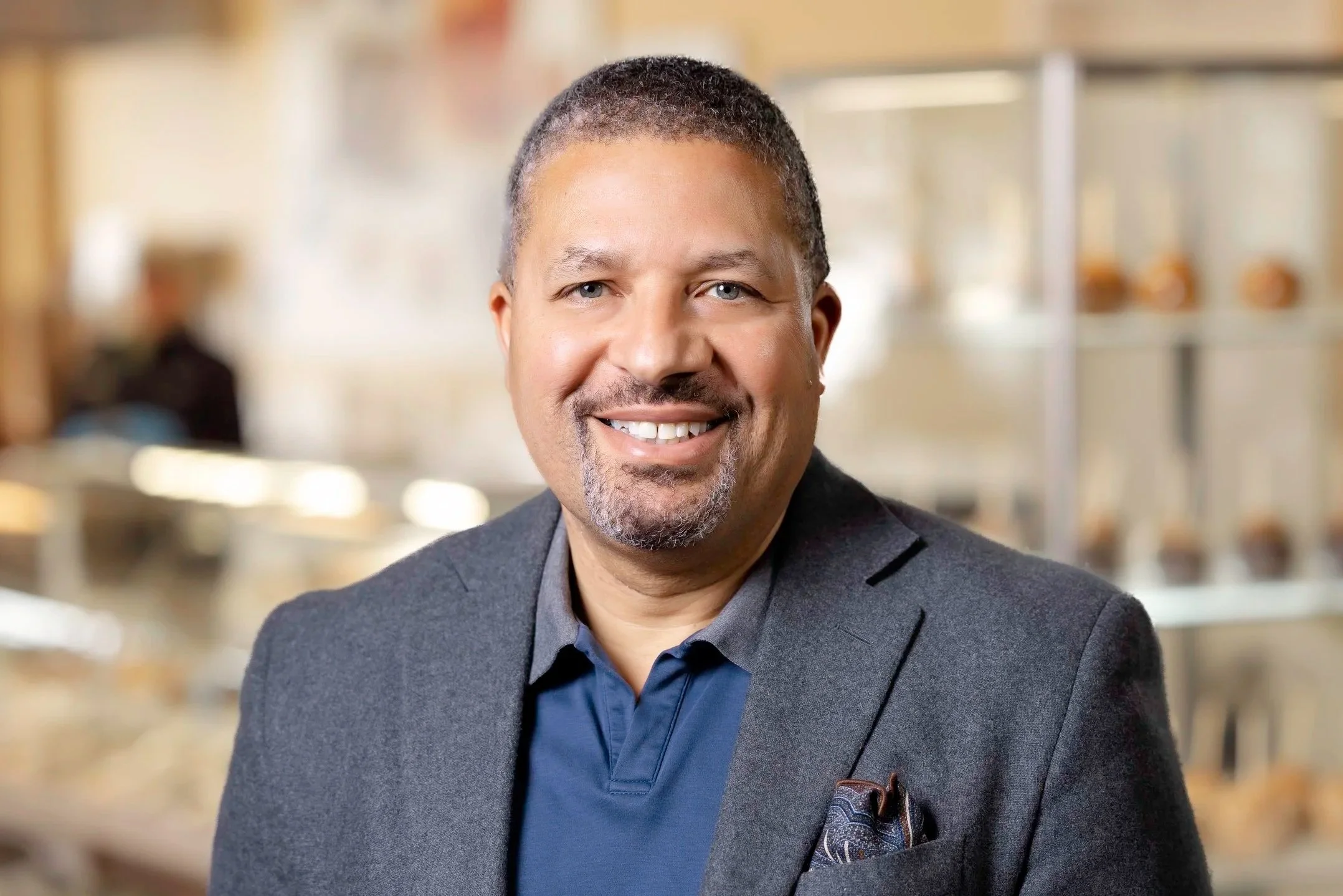 A smiling man with short hair and a goatee, wearing a dark blazer and a blue shirt in a bright indoor setting, possibly a cafe or bakery.