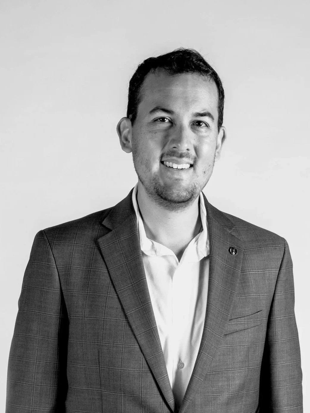 Black and white professional headshot of a man with short dark hair, smiling, wearing a checkered blazer and a white shirt, against a plain background.