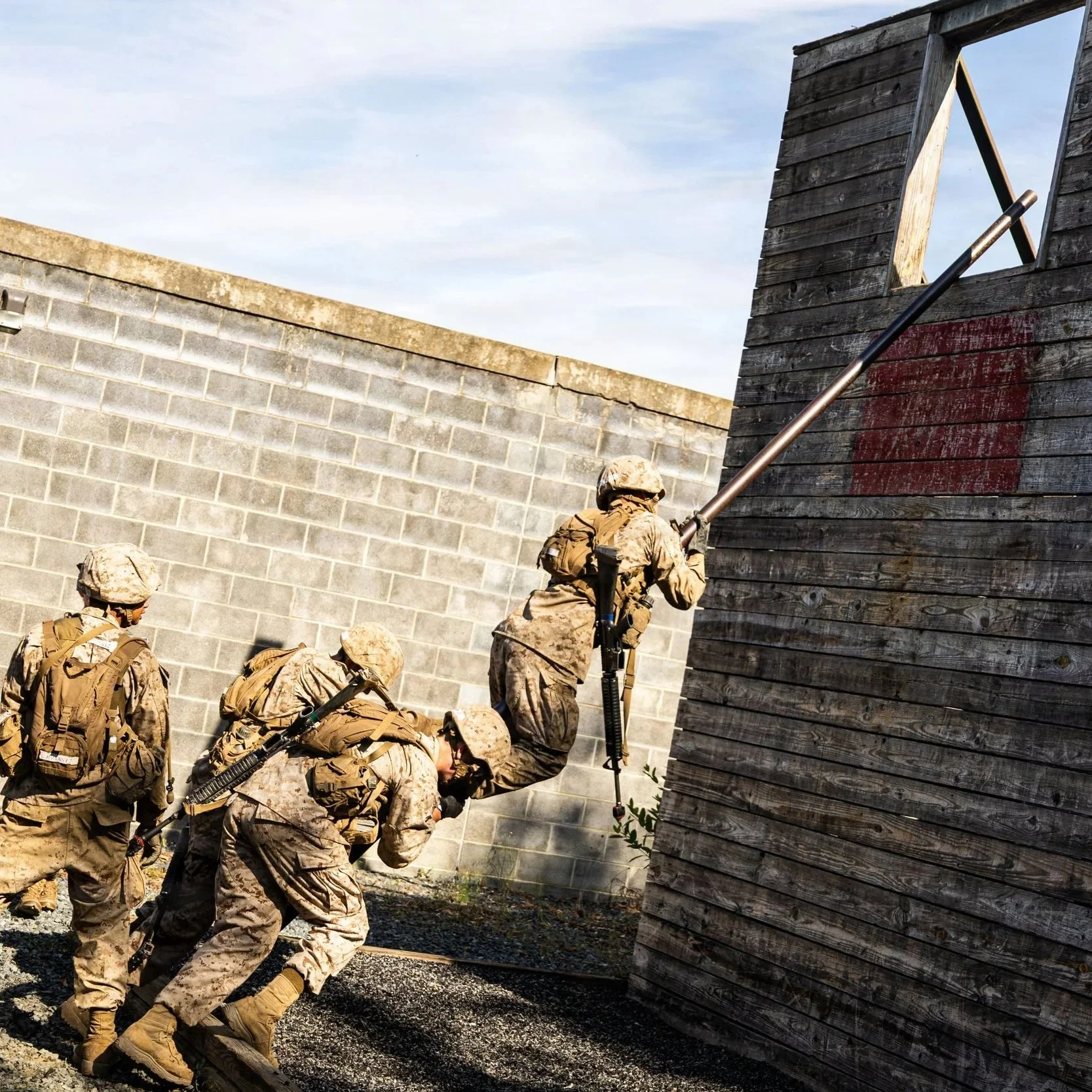 Soldiers in camouflage gear working together to climb a poll over a wall at a training exercise.