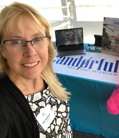 A woman with blonde hair, glasses, and a black and white patterned shirt smiling for a selfie at a booth with a laptop and informational materials.