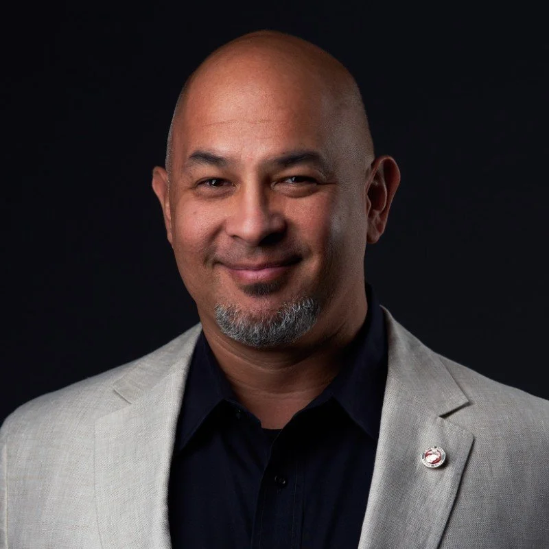 A portrait of a smiling man with a bald head, gray goatee, wearing a light gray blazer and black shirt, against a dark background.