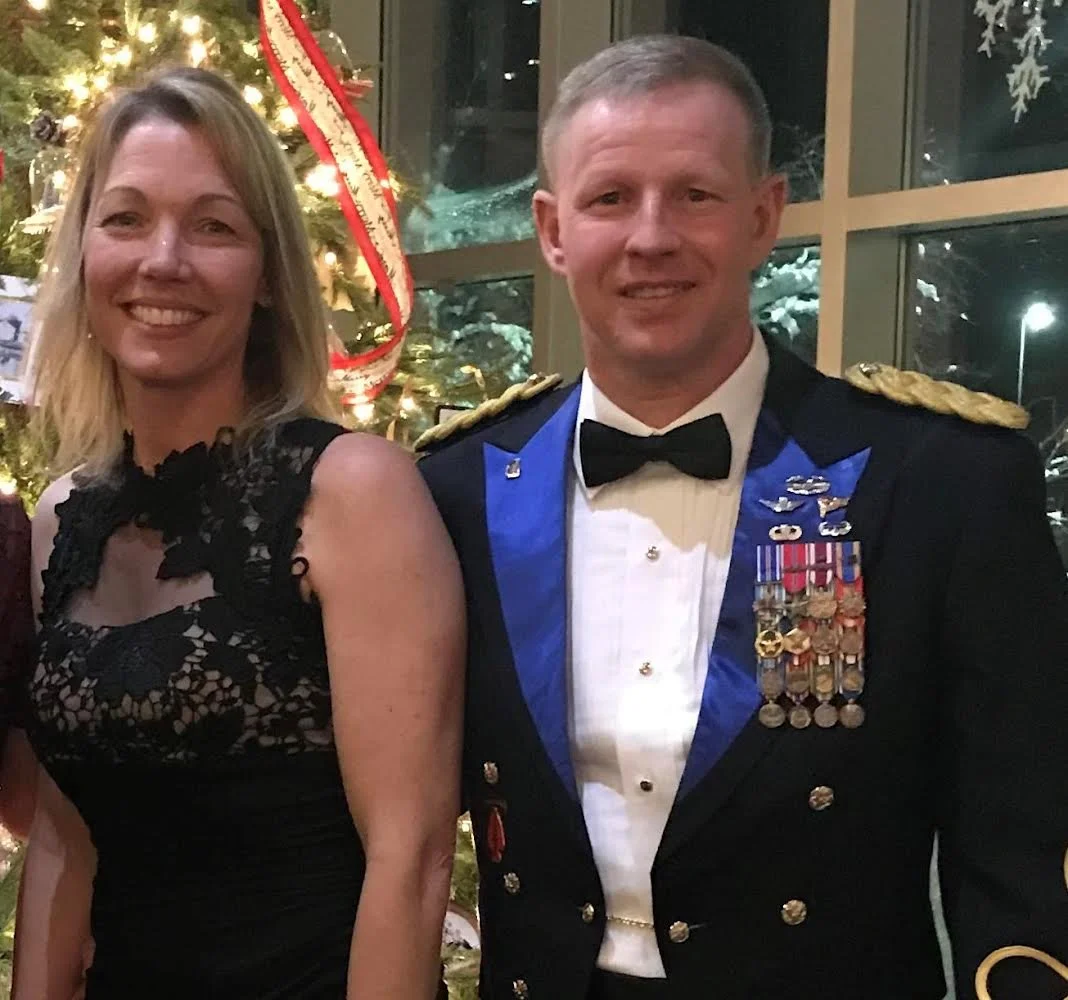 A smiling woman in a black lace dress standing next to a man in a military uniform with medals in front of a decorated Christmas tree.