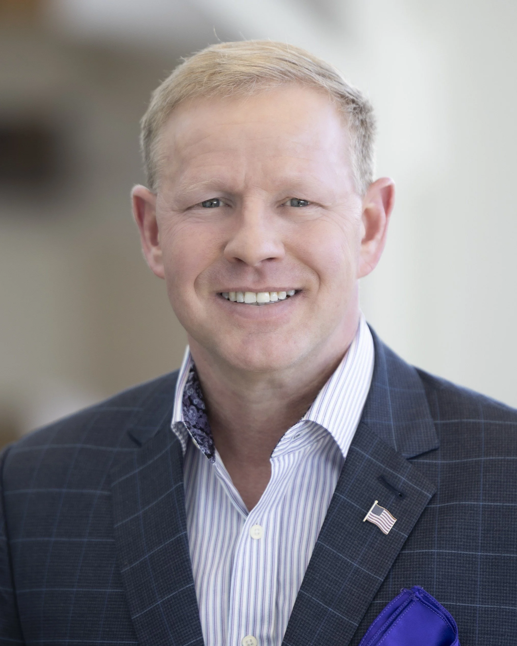 A man in a suit with a striped shirt smiling, wearing a small American flag pin on his lapel.