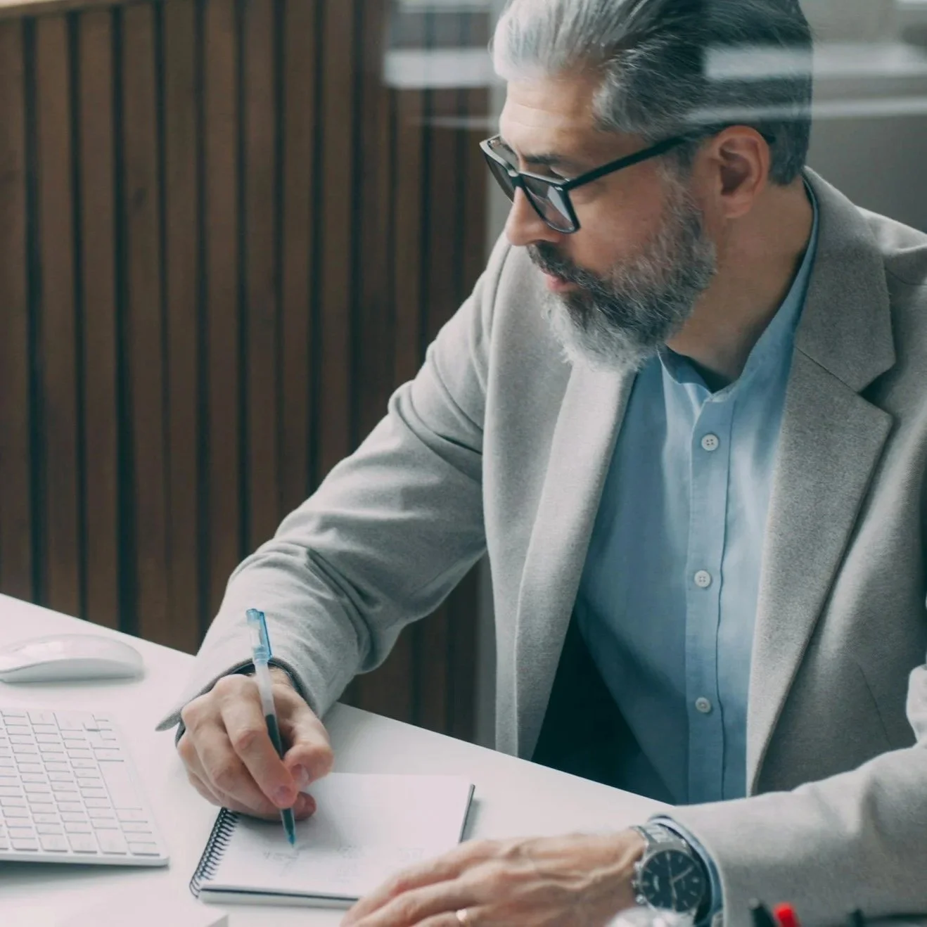 A man with gray hair and beard wearing glasses, a light gray blazer, and a blue shirt, sitting at a desk writing in a notebook with a pen, in an office setting.
