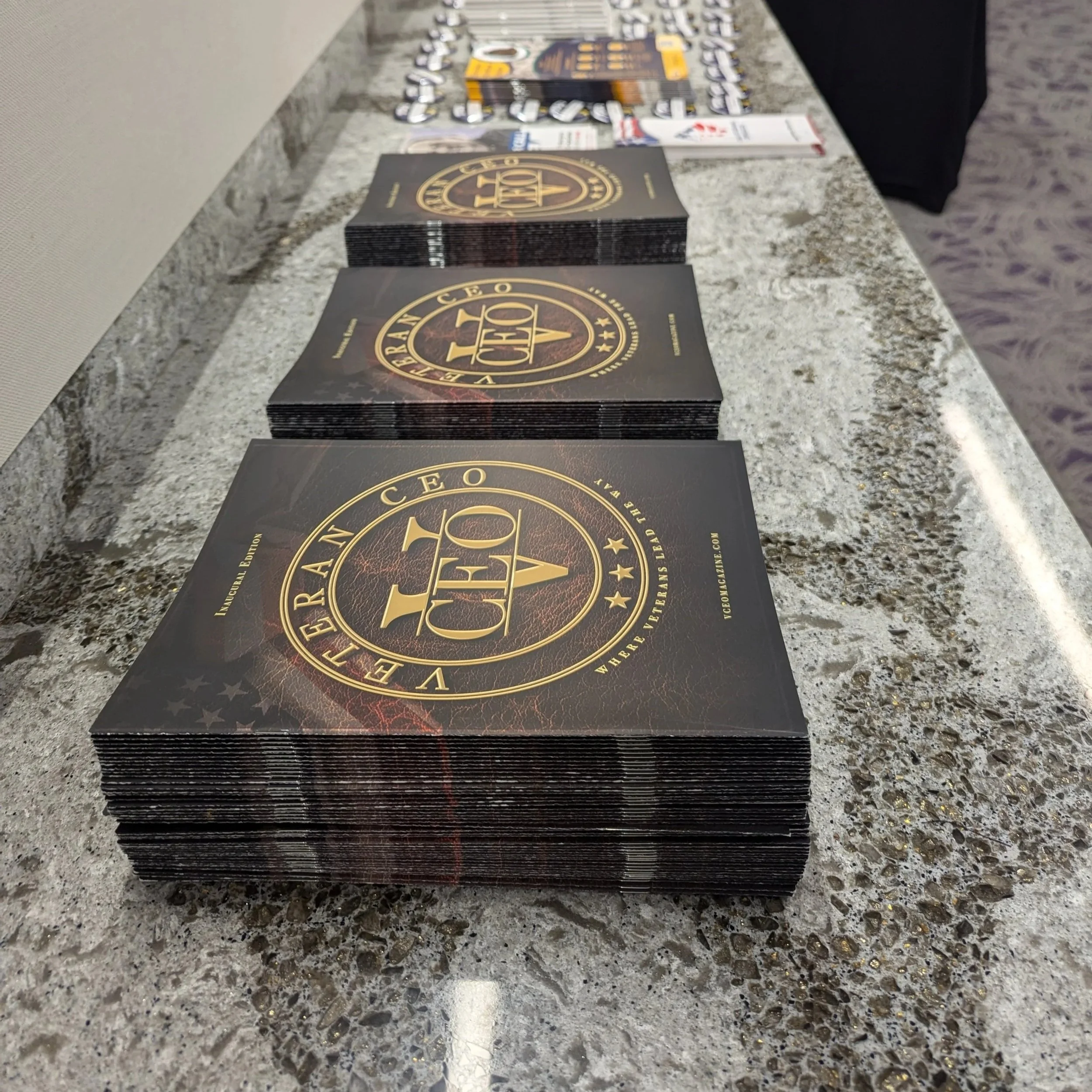 Stacks of black and gold brochures or booklets titled 'VETERAN CEO MAGAZINE - WHERE VETERANS LEAD THE WAY are arranged on a speckled gray countertop.