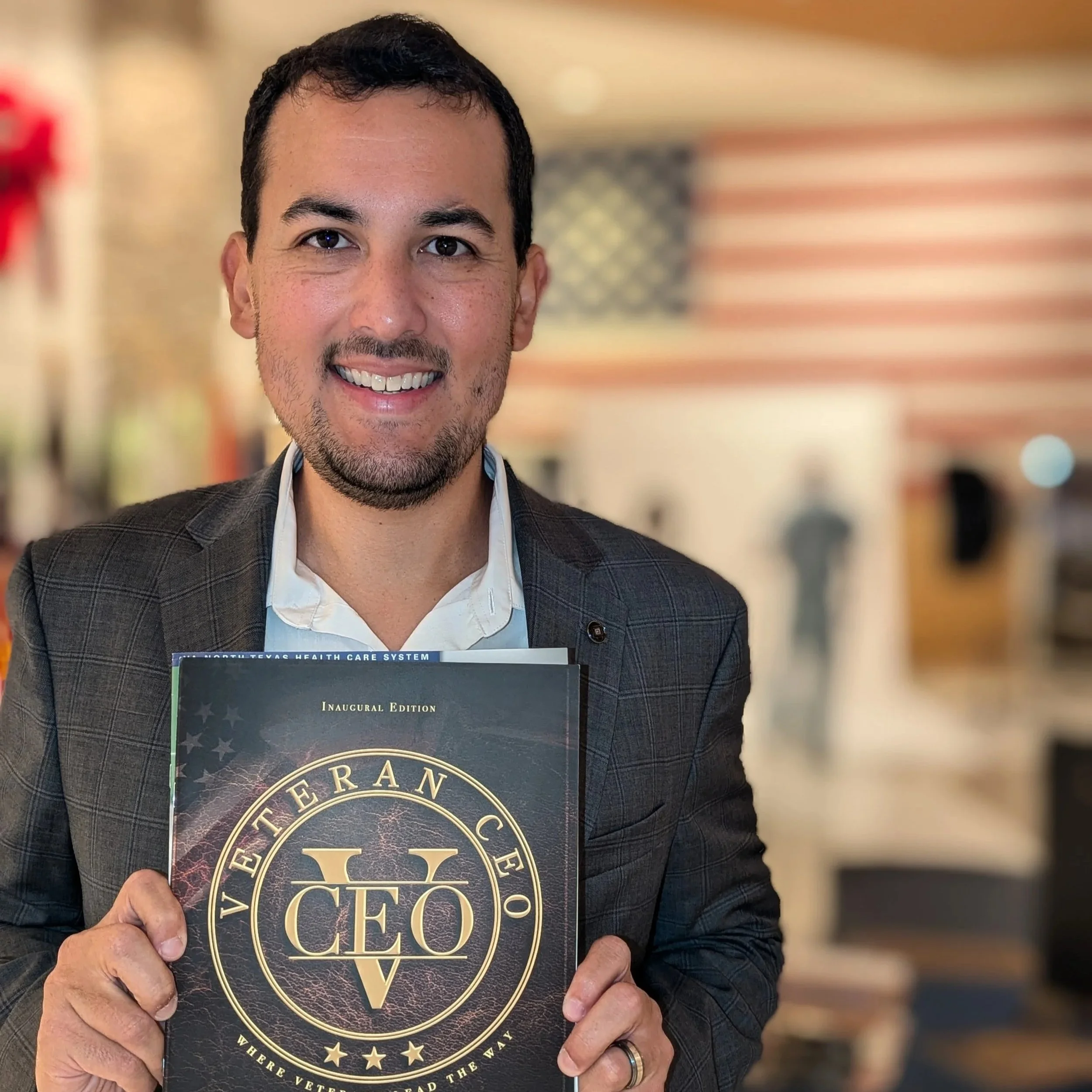 A man in a suit holds a copy of Veteran CEO Magazine with a large gold seal on the cover. The background is blurred, with an American flag visible and warm colors.