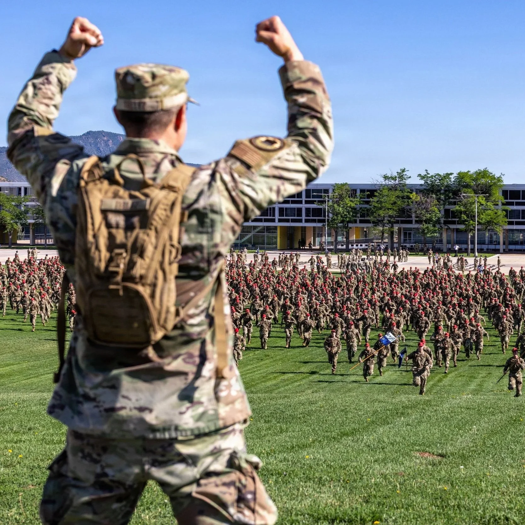 A soldier in camouflage military uniform and backpack raises fists in a field during a military gathering or ceremony. In the background, a large group of soldiers in similar uniforms march or stand in formation on a grassy area with trees, a modern building, and mountains under a clear blue sky.