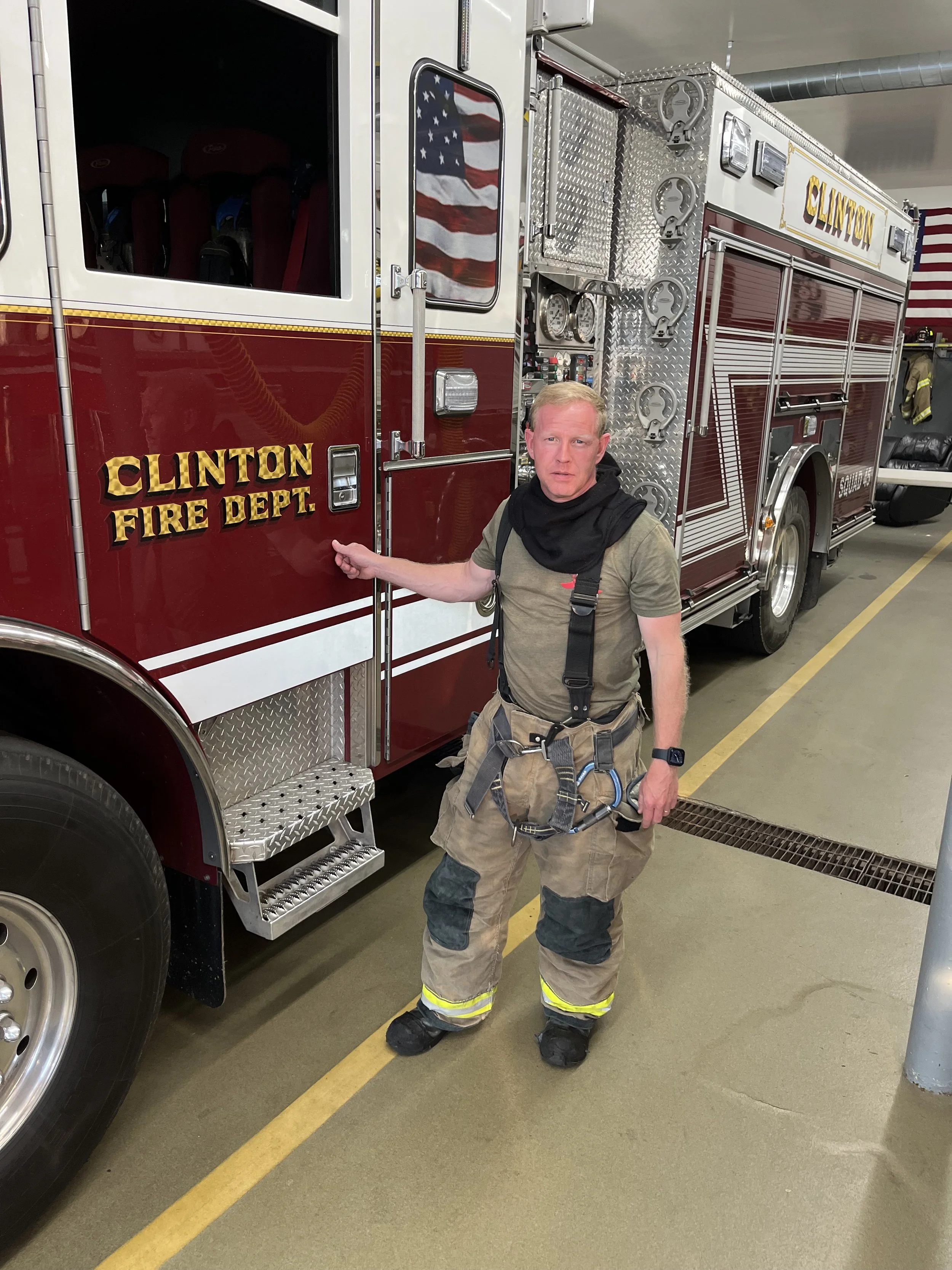 A firefighter in uniform standing next to a red Clinton Fire Department fire truck, pointing at the truck with one hand.