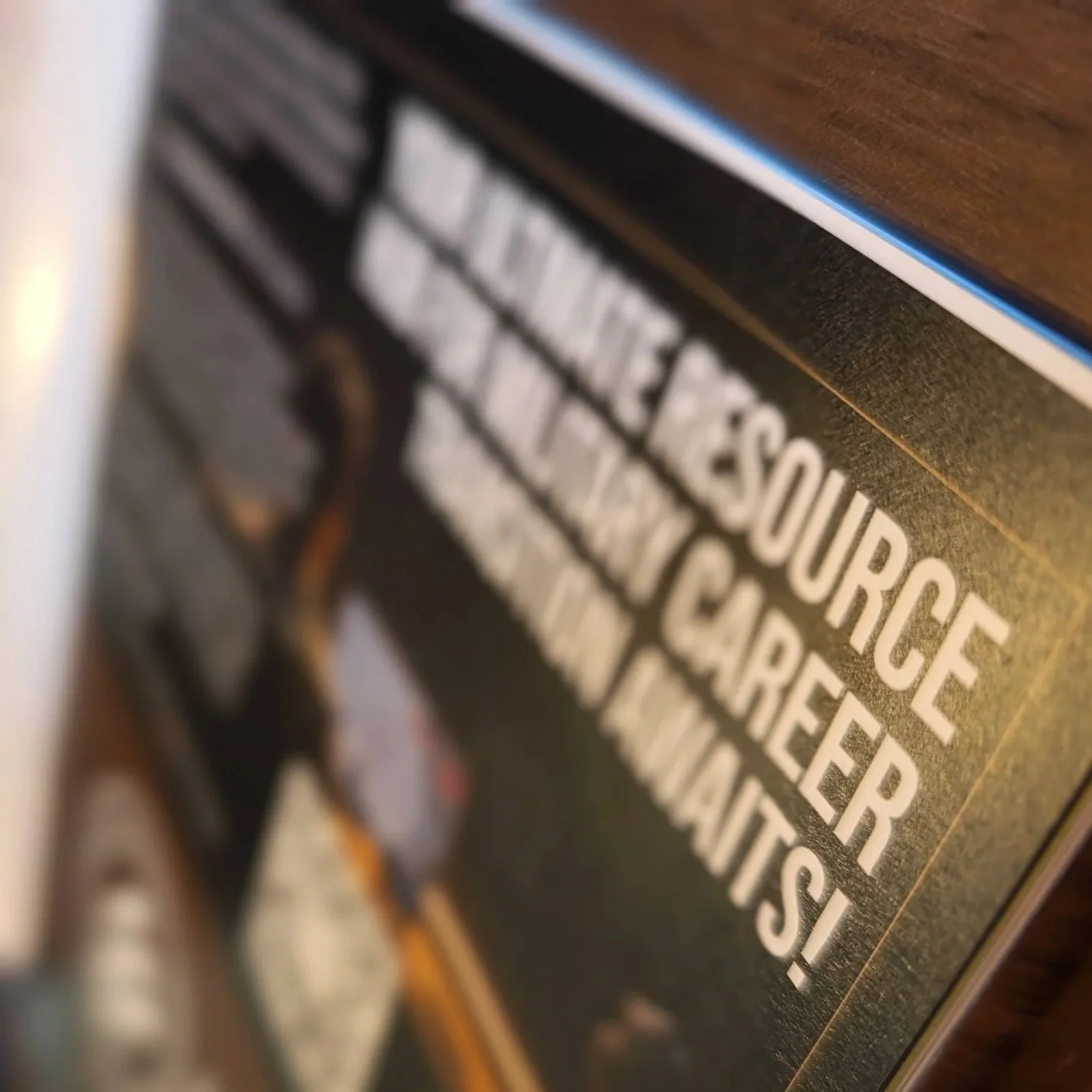 Close-up of a black page with white text reading 'RESOURCE, CAREER AWAITS!'. The text is mounted on a textured surface, possibly a wall or board, and a copper- or bronze-colored object with wires is partially visible on the left side of the image.