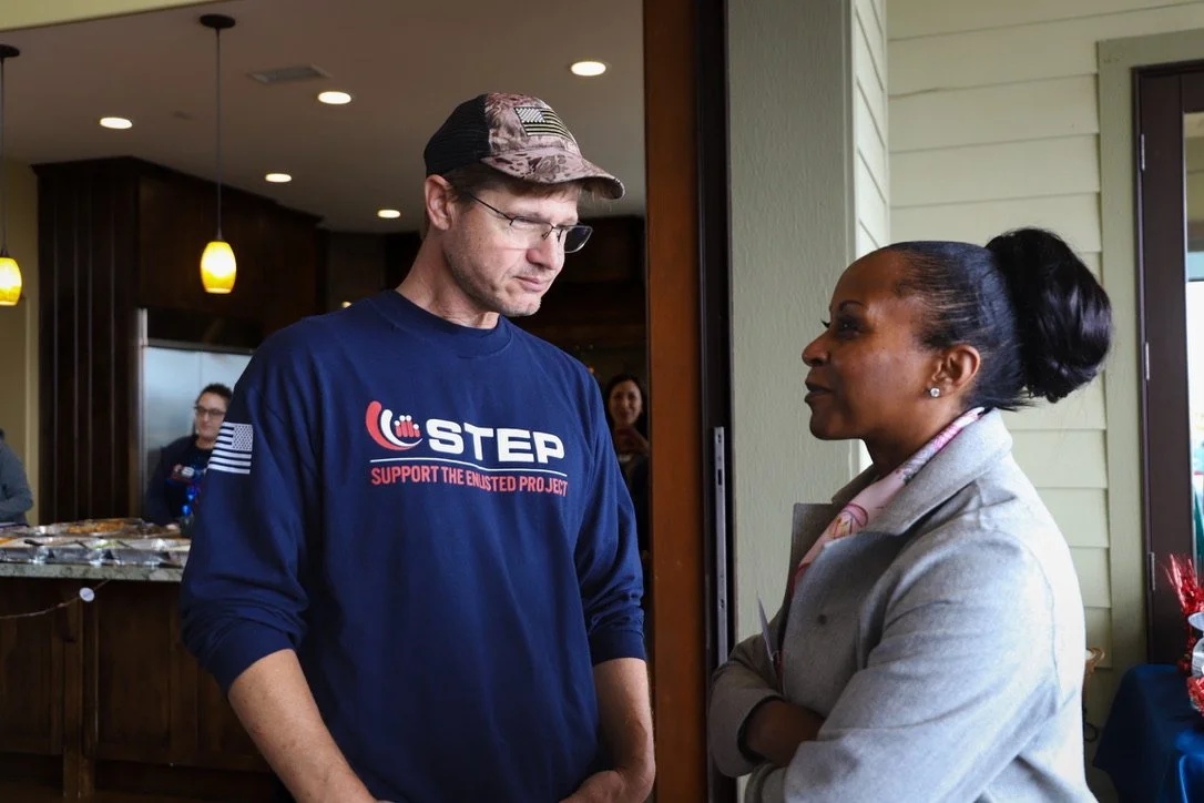A man wearing a blue sweatshirt with support for the enlisted project, a camouflage cap, and glasses, standing with arms crossed, conversing with a woman who has her arms crossed and is wearing a light gray coat, in an indoor setting near a door.