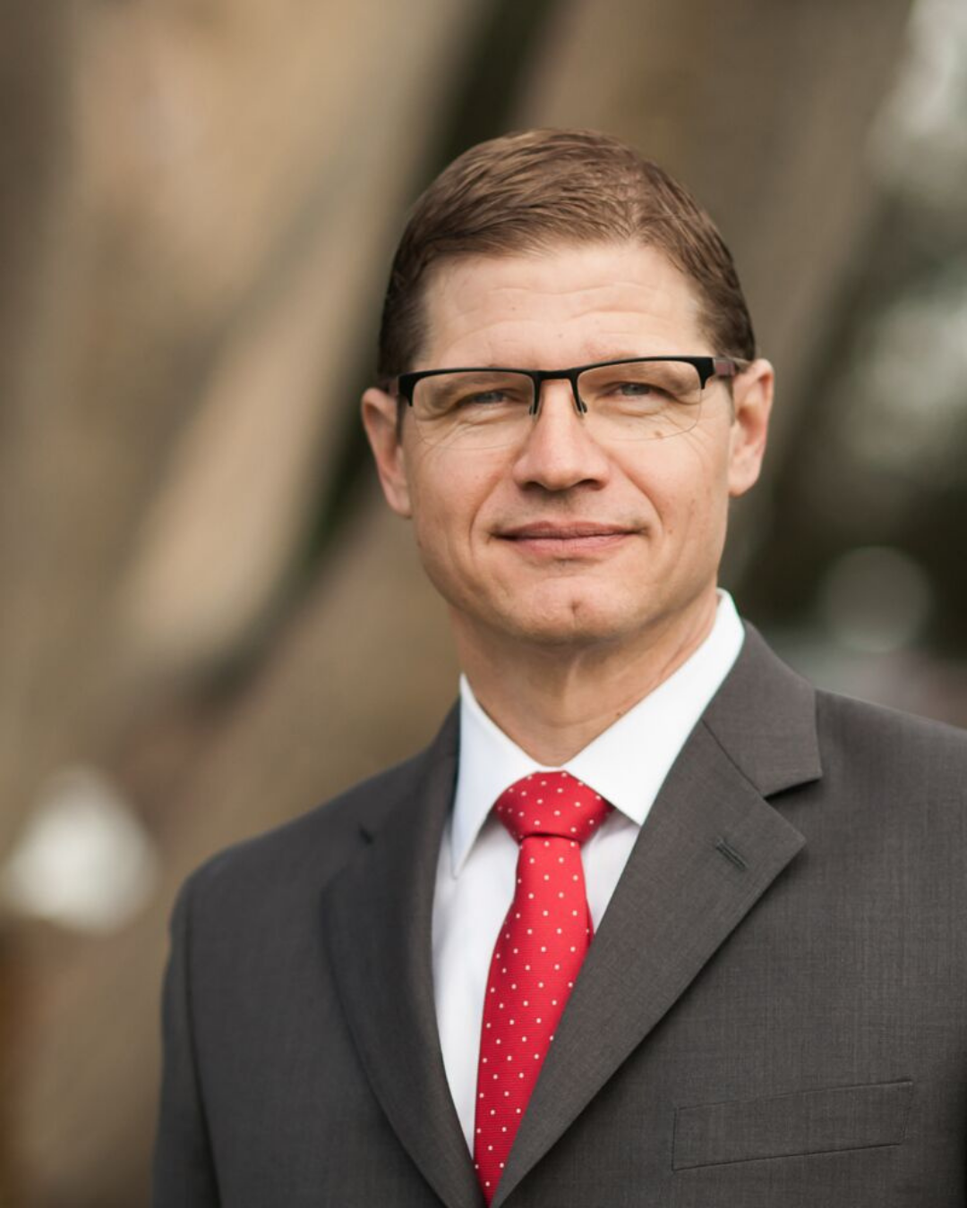 A man in a gray suit, white shirt, and red polka dot tie, wearing black glasses, standing outdoors with a blurred background.