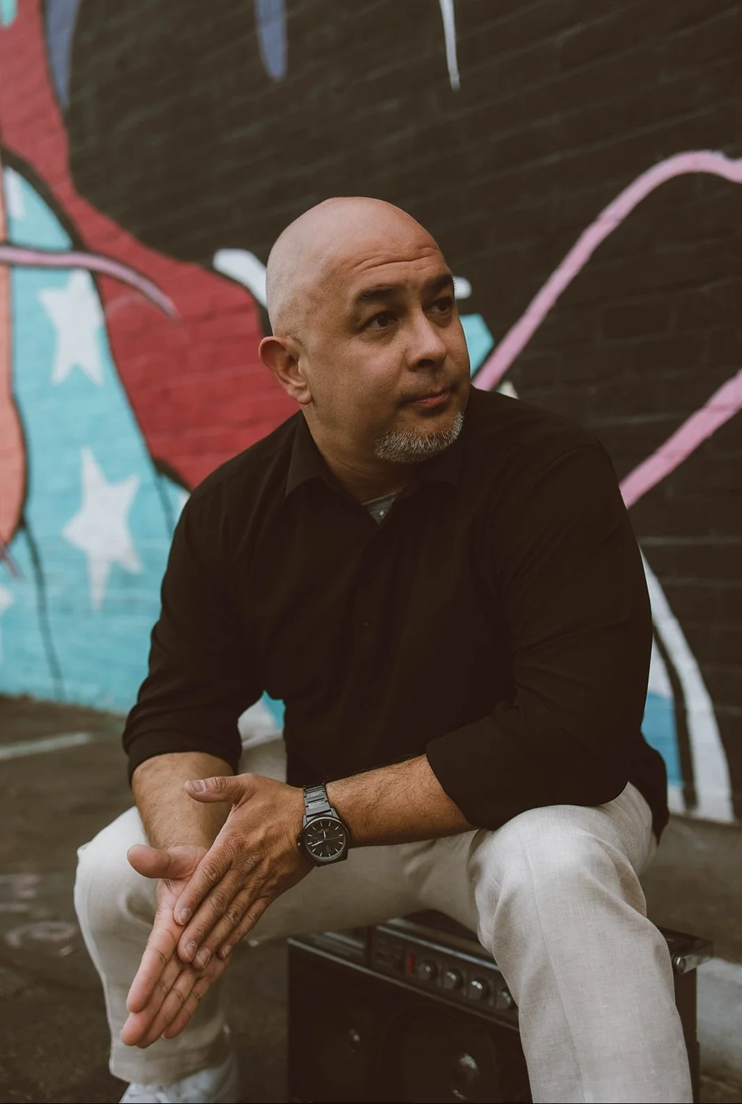 A man with a bald head and a goatee, wearing a black shirt, beige pants, and a black wristwatch, sitting on a small black box in front of a colorful graffiti wall.